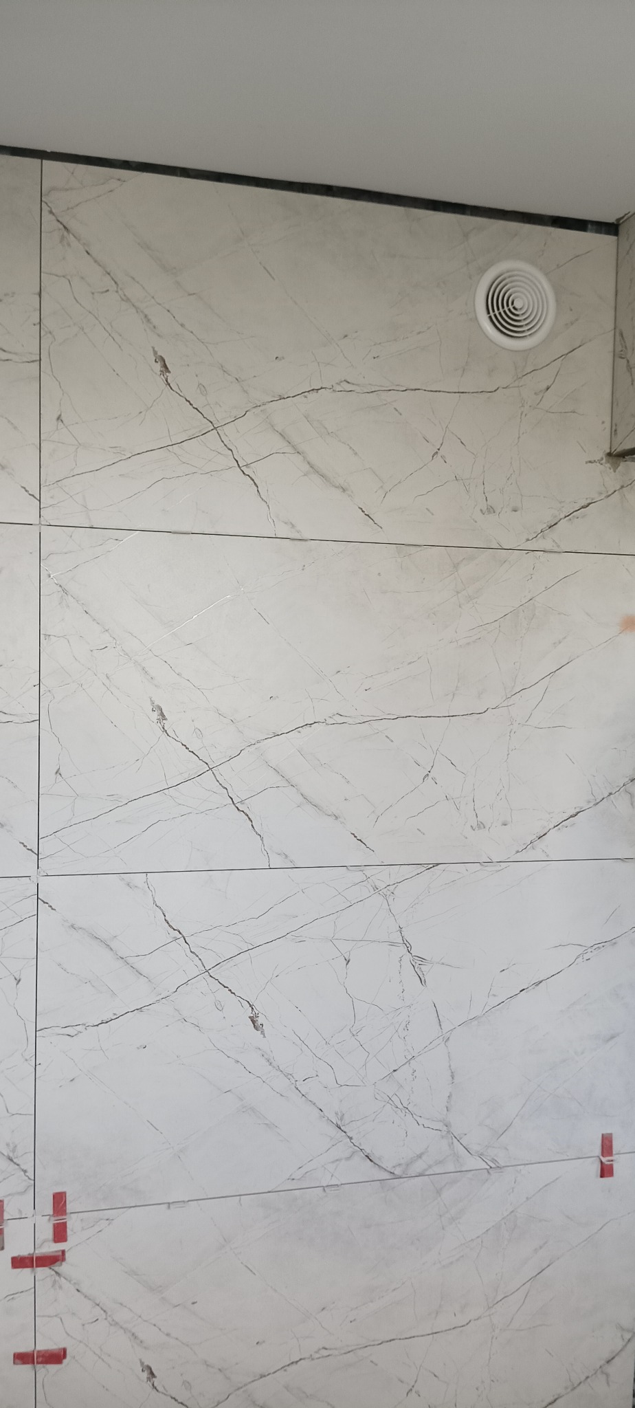 Ściana wyłożona białymi płytkami imitującymi marmur z widoczną kratką wentylacyjną i czerwonymi krzyżykami dystansowymi w dolnej części.