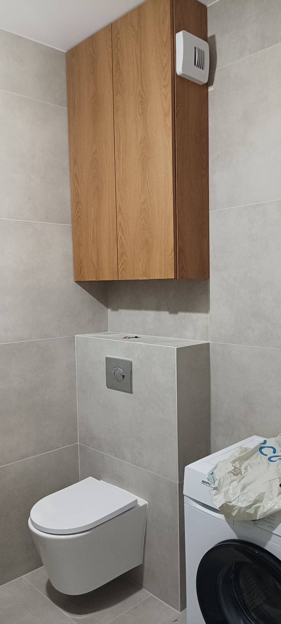 Nowoczesna, szara łazienka po remoncie: podwieszana toaleta, szafka z drewnianymi frontami, pralka. Minimalistyczny design, płytki w odcieniach szarości.