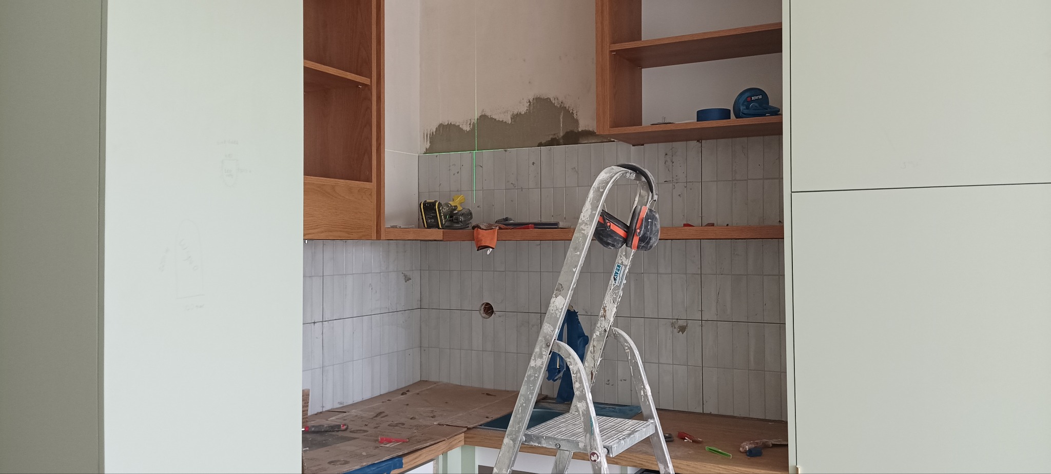 Remont kuchni: montaż płytek na ścianie, widoczna drabina, półki z drewna i narzędzia. Użycie lasera krzyżowego do poziomowania. Etap prac wykończeniowych.