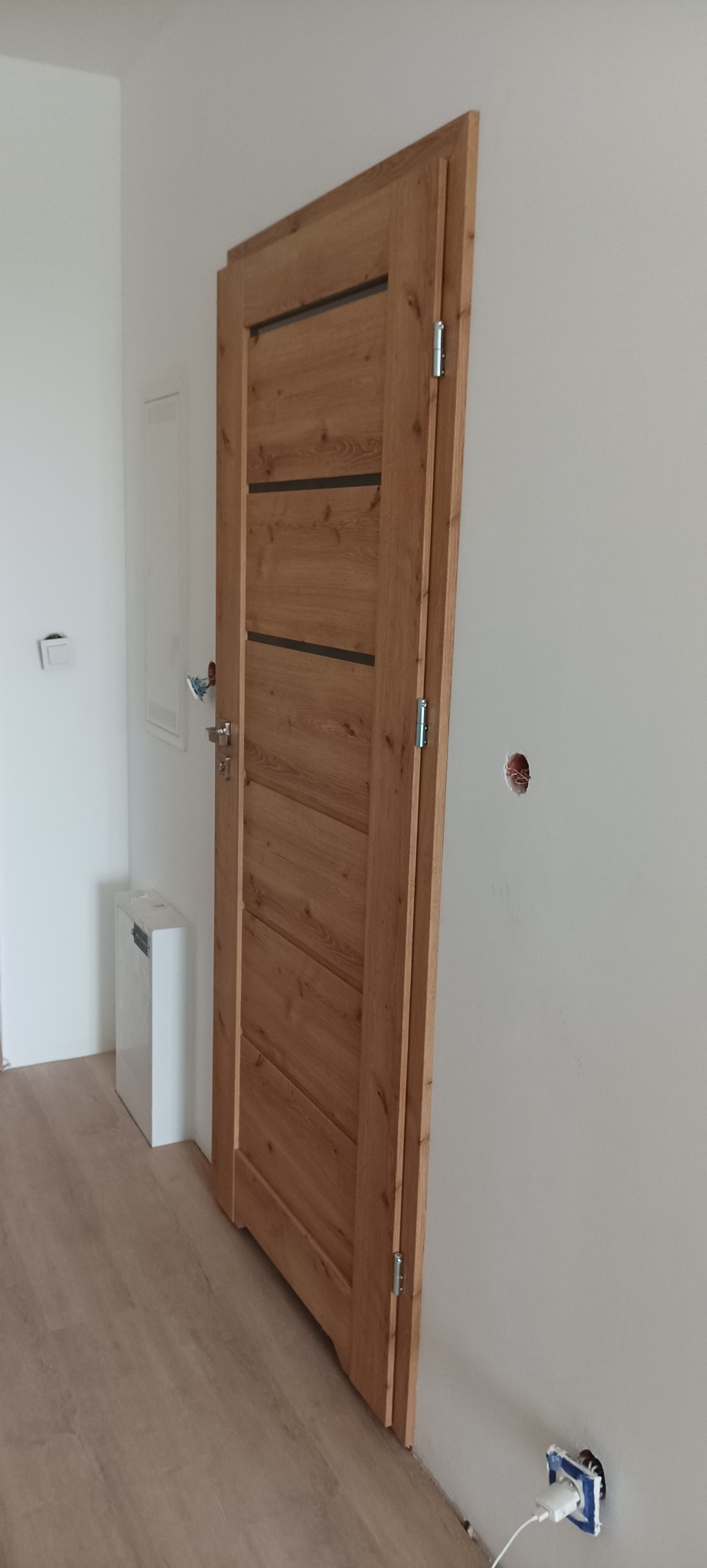 Zamontowane drewniane drzwi wewnętrzne z poziomymi, ciemnymi wstawkami, widoczne zawiasy i klamka. U dołu widoczny kontakt z ładowarką.