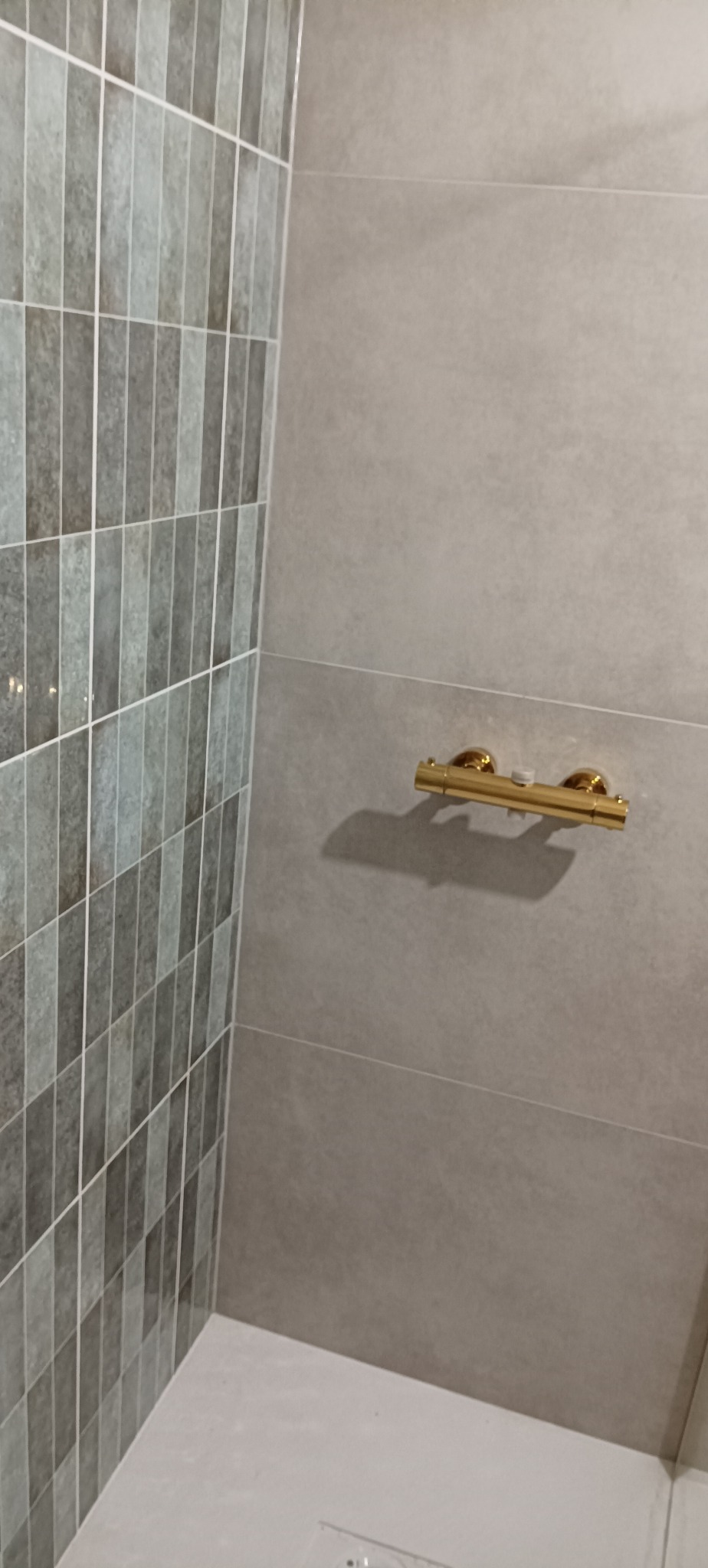 Wykończona łazienka z szarymi płytkami w pionowy wzór i złotą armaturą. Widoczny fragment brodzika. Minimalistyczny design, eleganckie wykończenie.