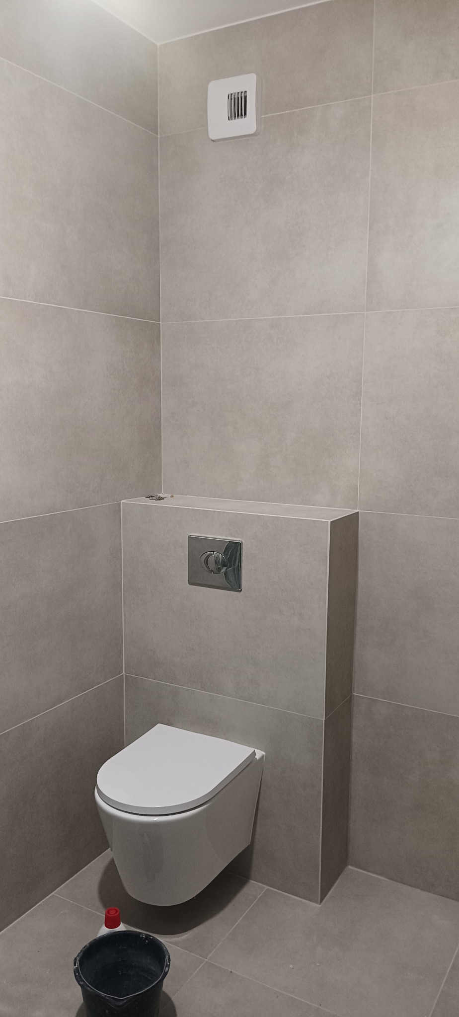 Szara łazienka po remoncie: podwieszana toaleta z chromowanym przyciskiem spłukiwania i wentylator. Na podłodze wiadro, a na ścianach duże, kwadratowe płytki.