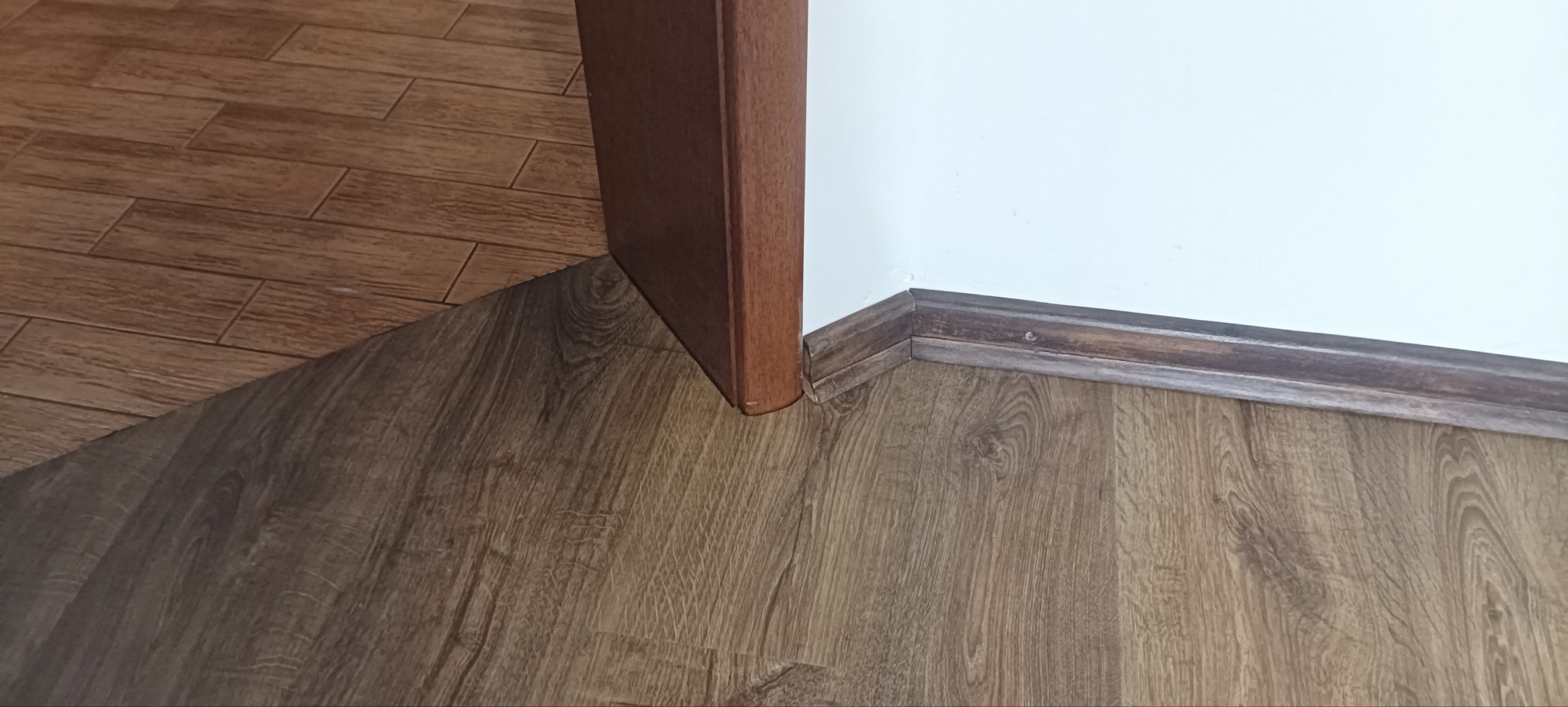 Fragment podłogi z paneli imitujących drewno, stykających się z białą ścianą i brązową listwą przypodłogową oraz fragmentem brązowej ościeżnicy drzwiowej.