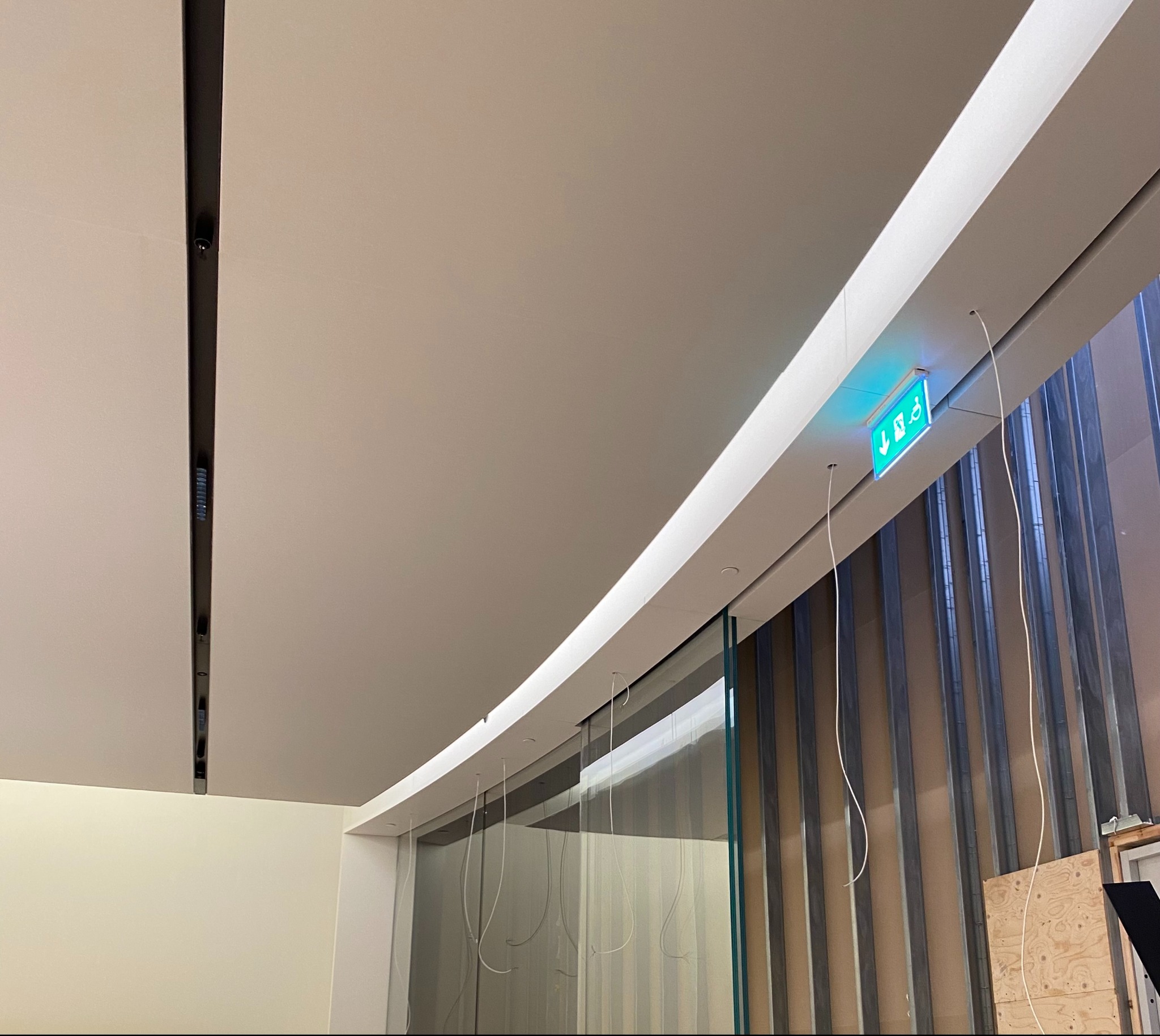 Sufit podwieszany z widoczną konstrukcją nośną i oświetleniem awaryjnym, w tle szklana ściana z metalowymi profilami.