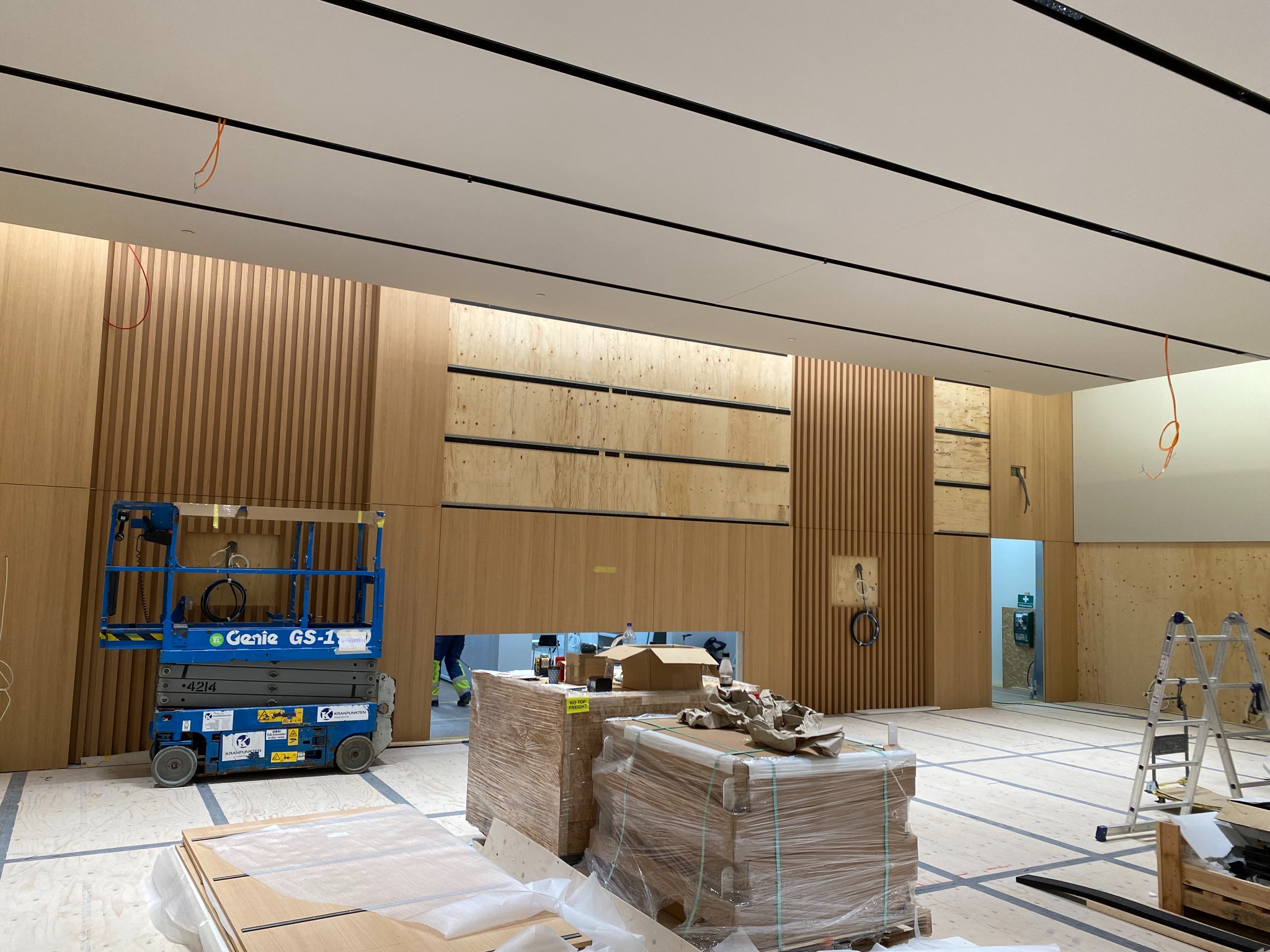 Wnętrze biura w trakcie prac wykończeniowych: widoczna podnośnik koszowy Genie, zabezpieczone folią palety z materiałami budowlanymi, drewniane panele ścienne i sufit z czarnymi listwami.