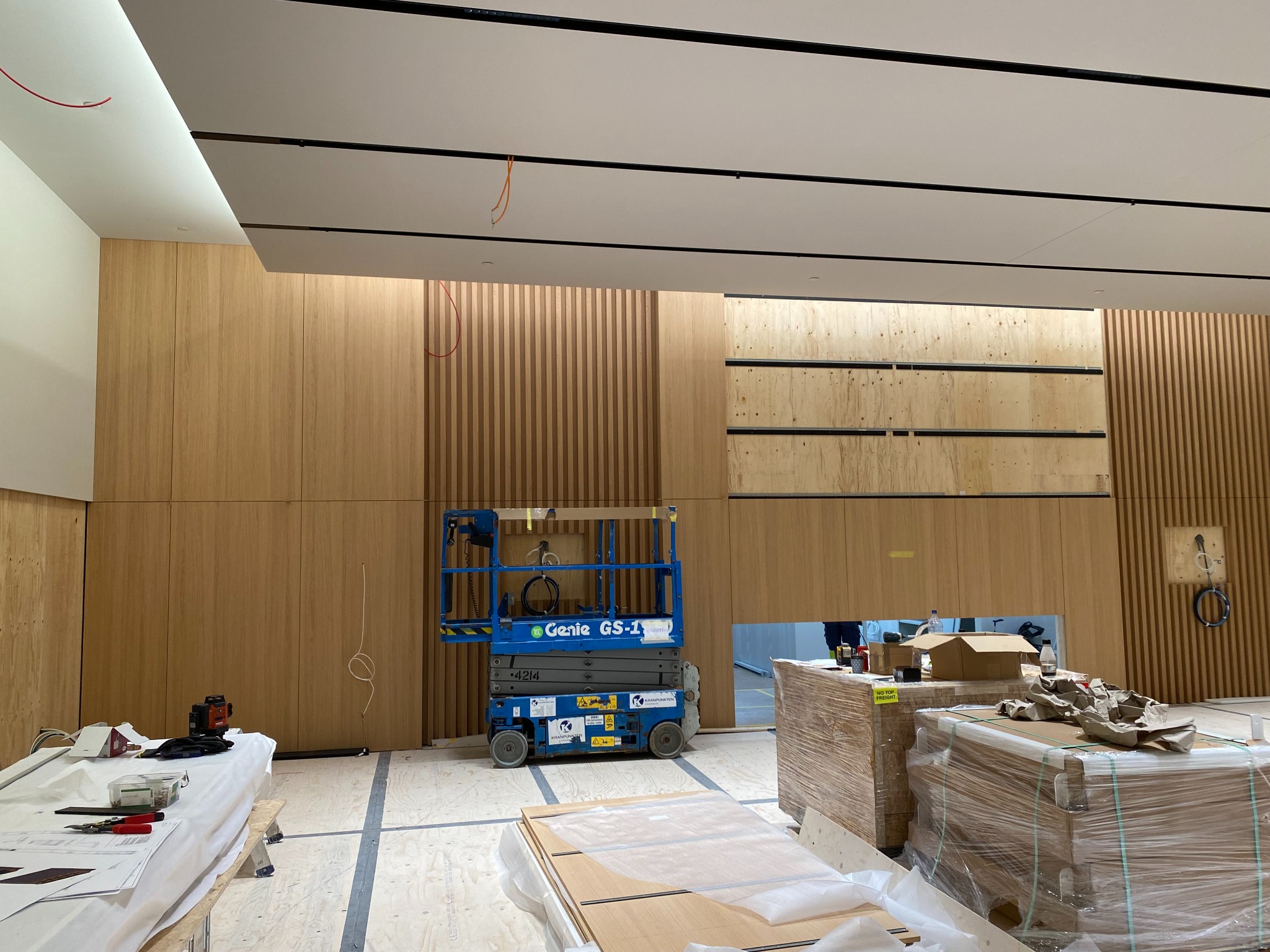 Wnętrze biura w trakcie prac wykończeniowych: widoczna podnośnik nożycowy Genie GS-1, drewniane panele ścienne, przygotowane elementy do montażu, instalacja oświetleniowa na suficie.