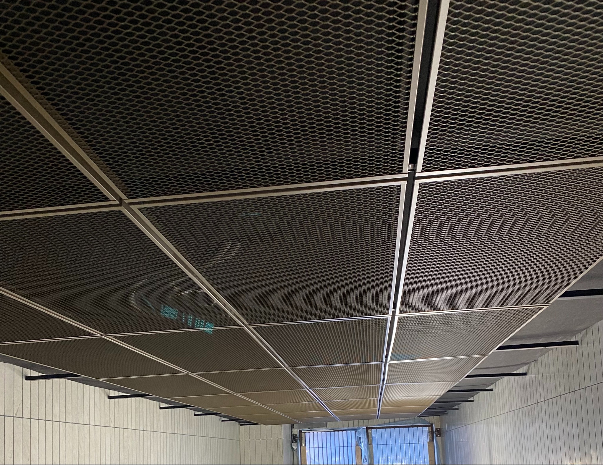 Rzut z dołu na metalowy sufit podwieszany w biurze, z widoczną siatką i ramami nośnymi, oświetlenie w tle.