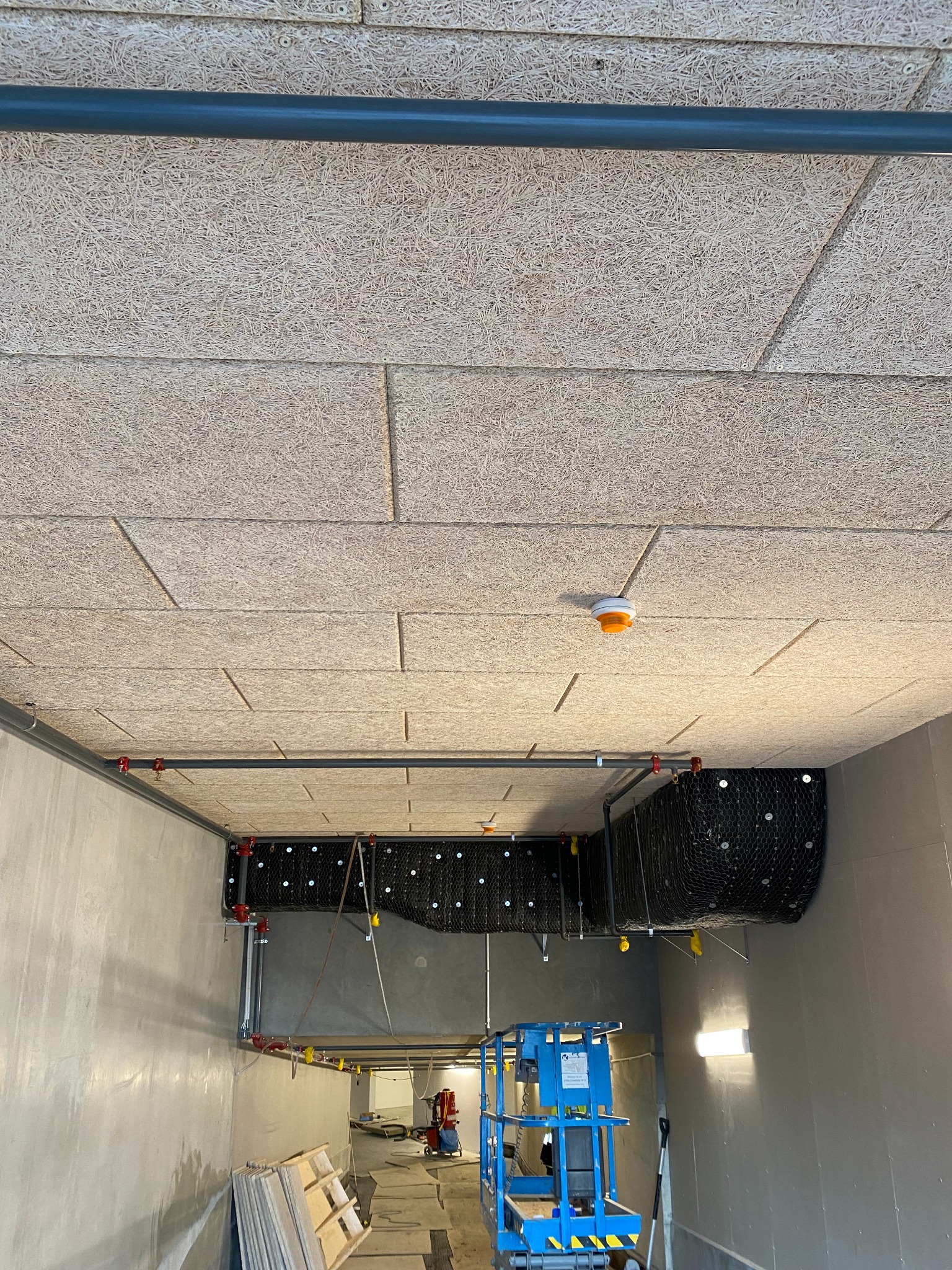 Montaż sufitu z paneli drewnopochodnych o strukturze włóknistej, widoczne instalacje pod sufitem, w tle podnośnik nożycowy i materiały budowlane.