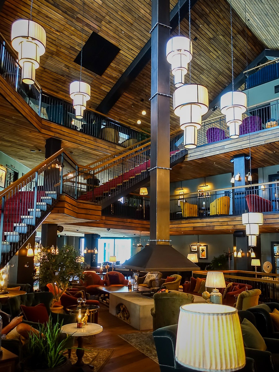 Przestronne wnętrze hotelu z drewnianym sufitem, schodami z czerwonym dywanem, wielopoziomowymi balkonami i centralnie umieszczonym kominkiem z okapem, oświetlone wiszącymi lampami w stylu art deco.