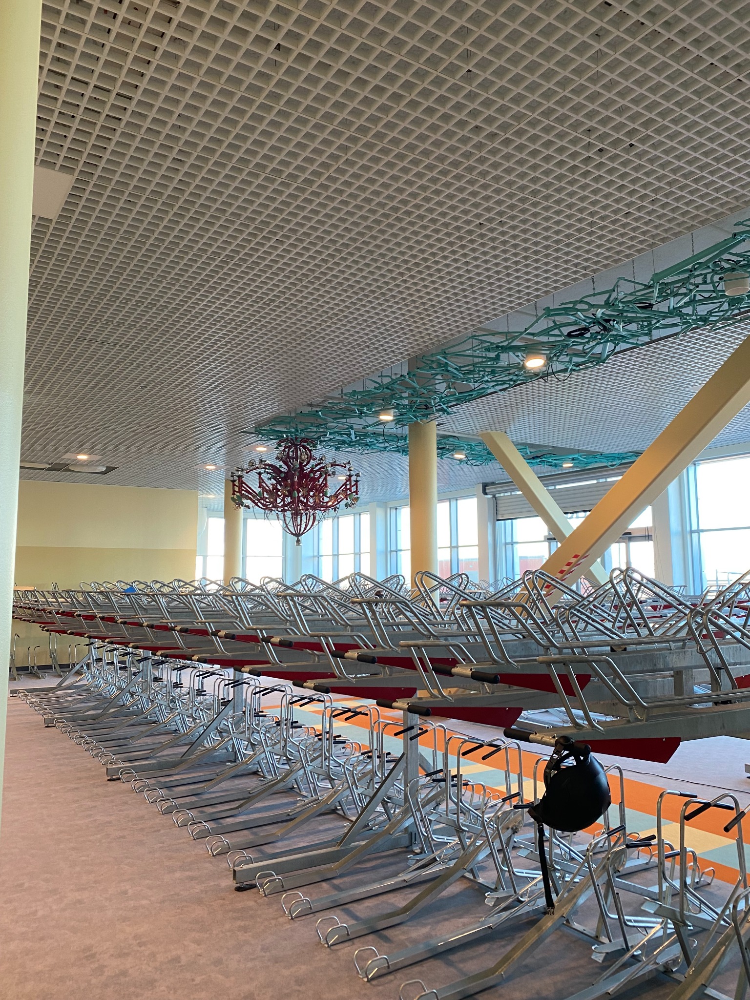 Rzędy pustych, metalowych stojaków na rowery w jasnym wnętrzu z kasetonowym sufitem i czerwonym żyrandolem, widoczny kask na jednym ze stojaków.