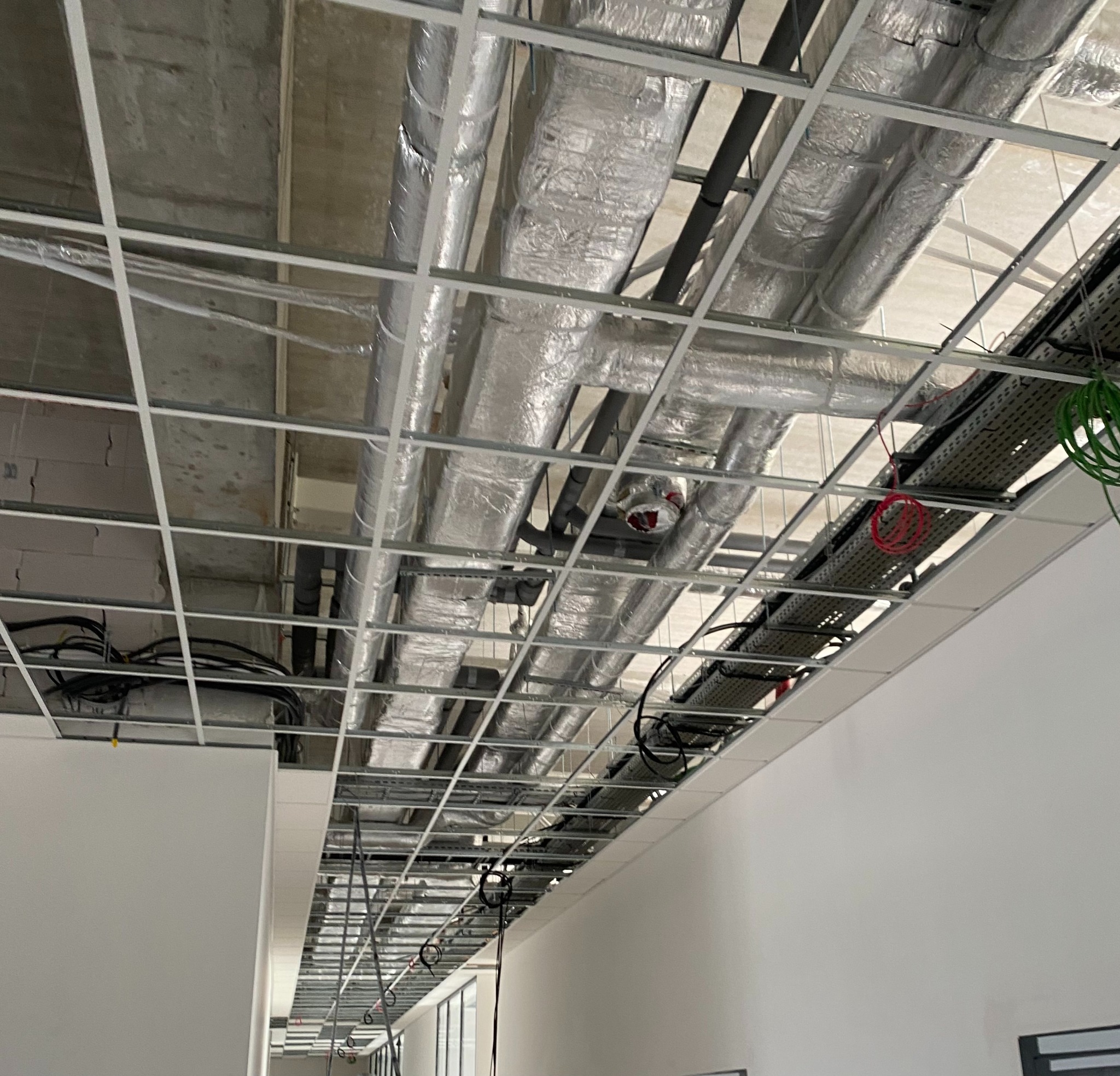 Widok sufitu podwieszanego w trakcie montażu, widoczne przewody wentylacyjne w izolacji, kable elektryczne i elementy konstrukcyjne kratownicy, ściany pomalowane na biało, oświetlenie w trakcie...