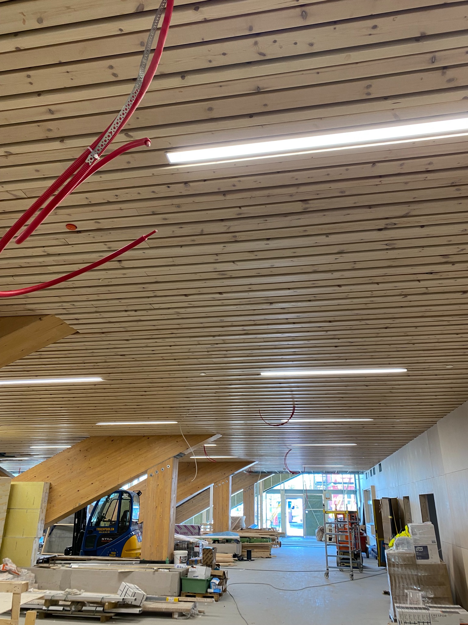 Drewniany sufit lamelowy z widocznymi instalacjami elektrycznymi w rurkach PCV, fragment konstrukcji drewnianej i wózek widłowy w tle, w trakcie prac wykończeniowych.
