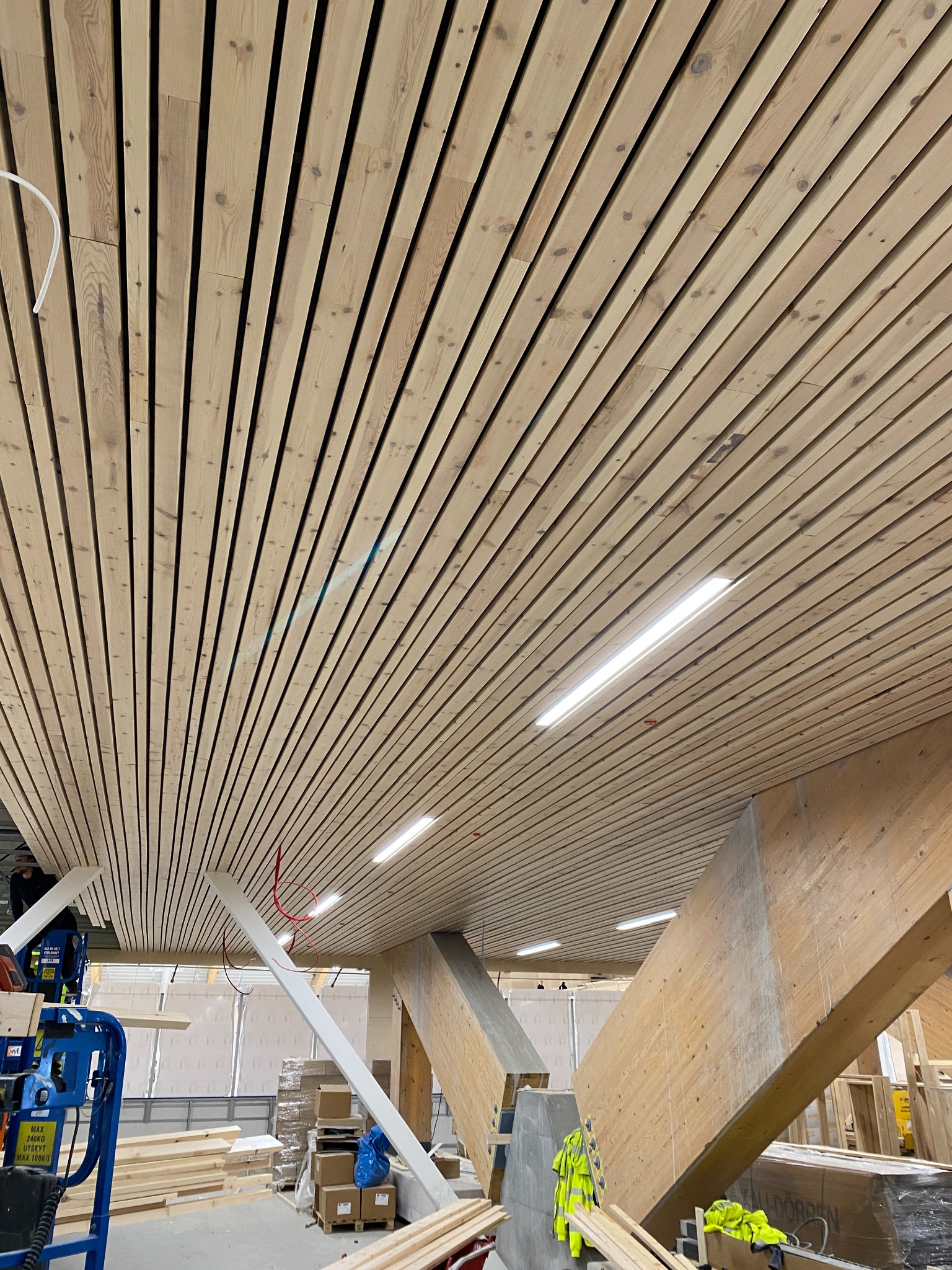 Drewniany sufit lamelowy z oświetleniem LED, widoczne elementy konstrukcyjne i maszyna budowlana w tle.