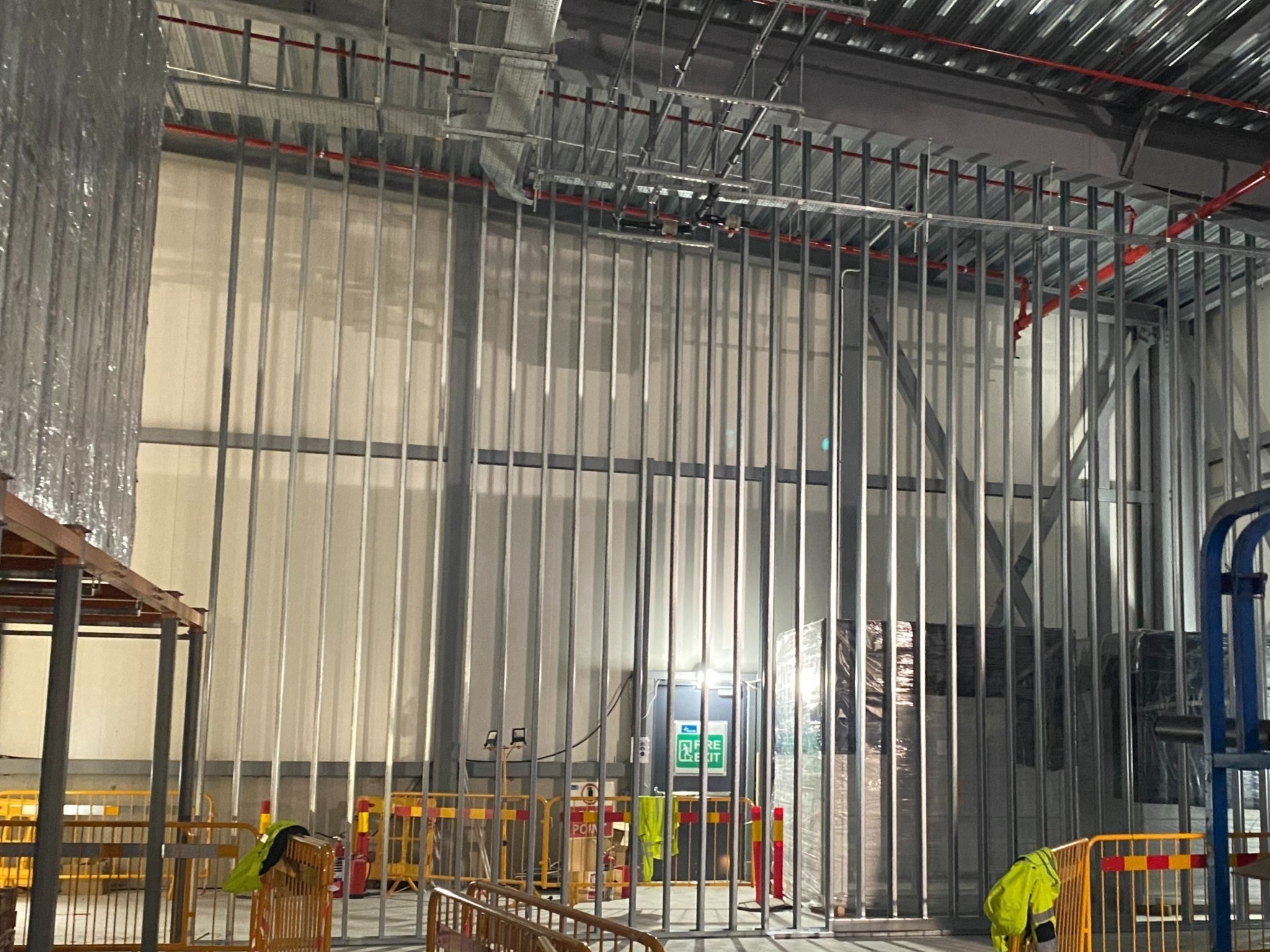 Montaż metalowej konstrukcji szkieletowej ścian działowych w hali magazynowej, widoczne instalacje pod sufitem i tymczasowe zabezpieczenia BHP