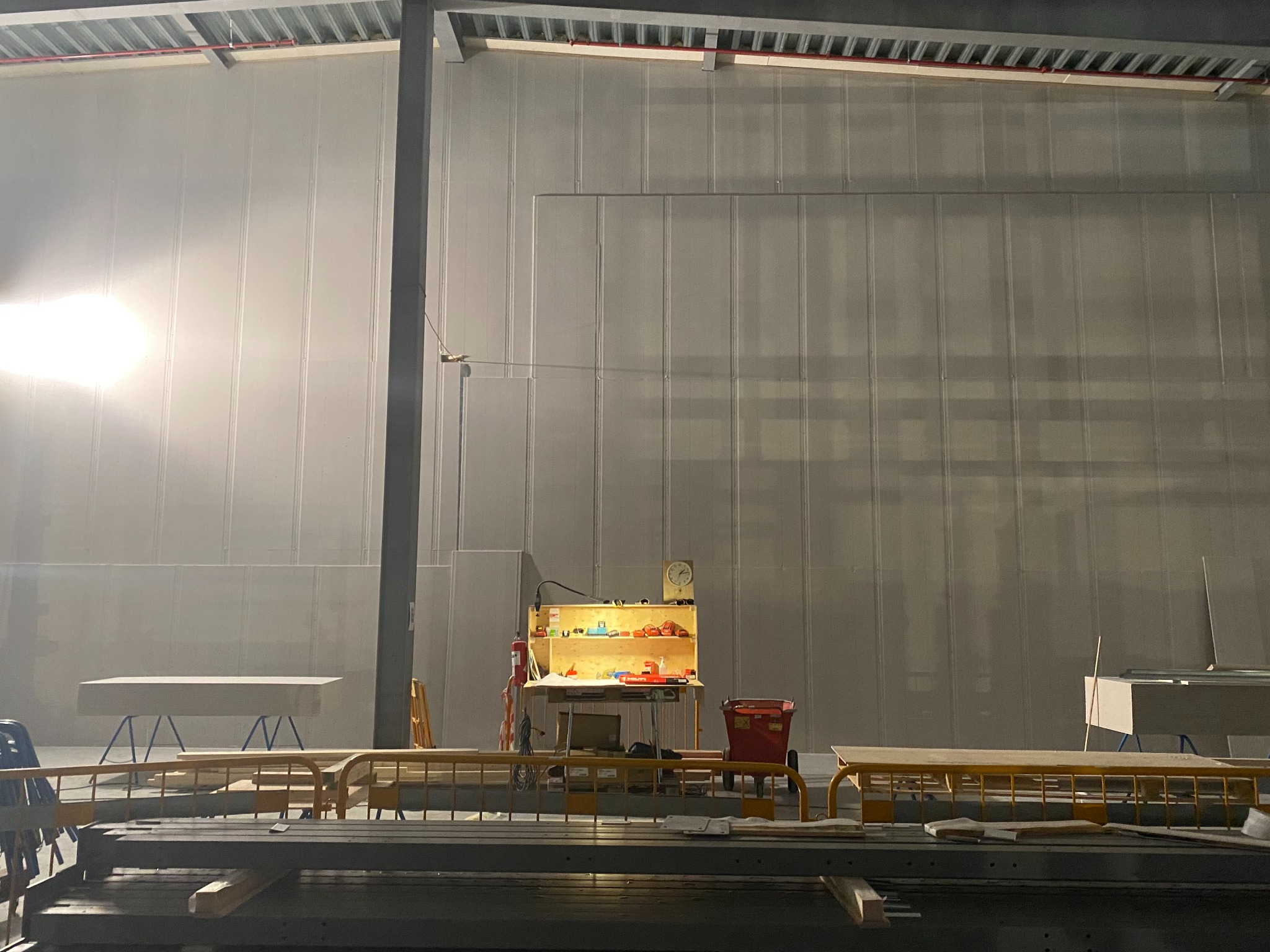 Wnętrze hali w trakcie prac wykończeniowych, widoczna ściana z płyt warstwowych, prowizoryczny warsztat z narzędziami i materiałami budowlanymi podświetlony lampą, oraz elementy konstrukcji...