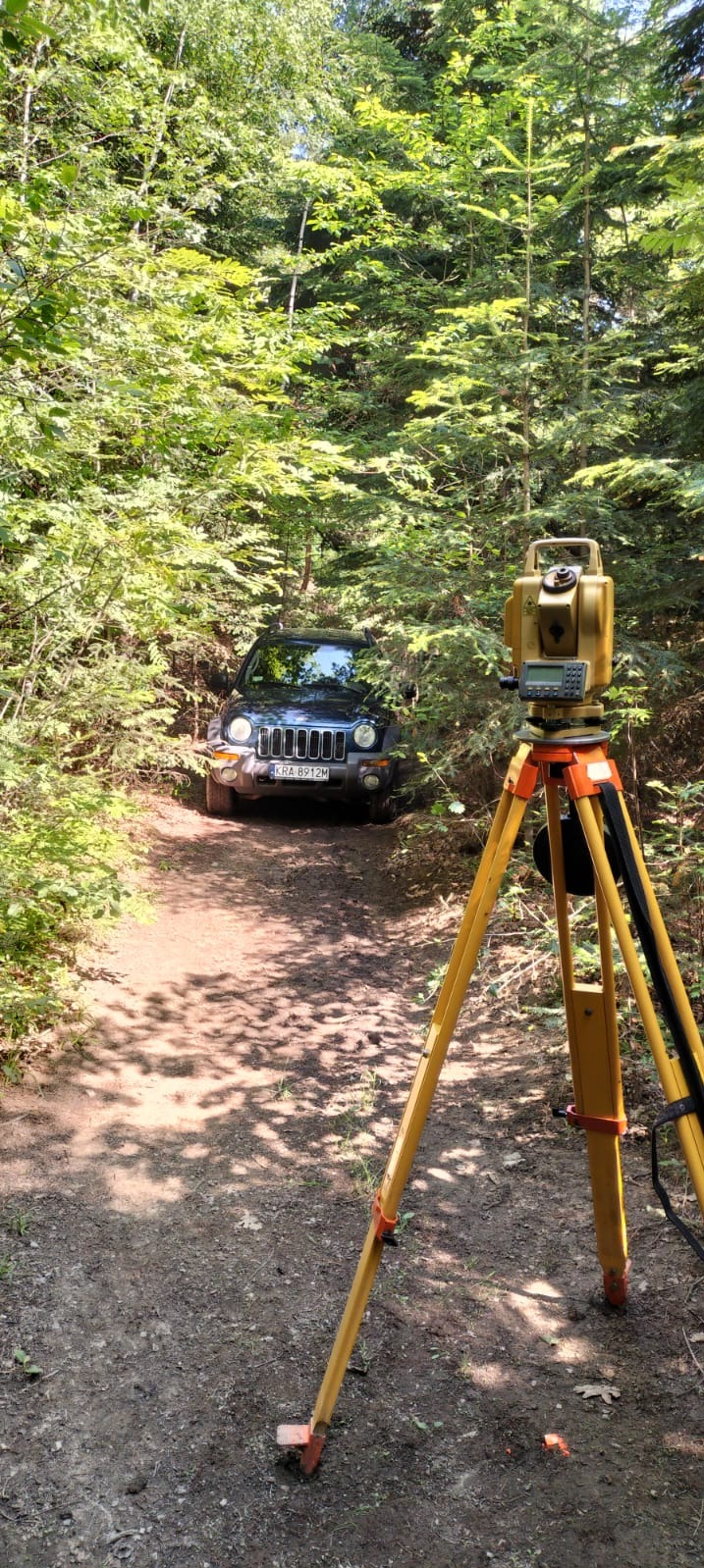 Geodezyjny tachimetr na trójnogu ustawiony na leśnej drodze, w tle ciemny samochód terenowy, drzewa i krzewy.
