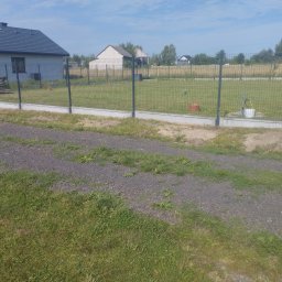 Kuba mąkosa - Nowoczesne ogrodzenie panelowe z podmurówką betonową na tle zieleni i zabudowań. Widoczne słupki i siatka ogrodzeniowa. Jasne, słoneczne oświetlenie.