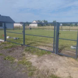 Szara, metalowa brama panelowa w Radomiu, z widoczną furtką i fragmentem ogrodzenia. W tle dom i trawnik, brama otwarta na oścież, słoneczny dzień.