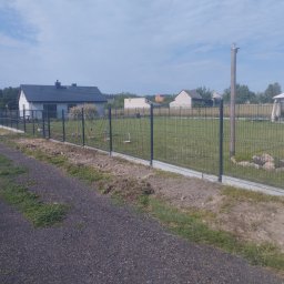 Kuba mąkosa - Nowo postawione ogrodzenie panelowe z podmurówką na tle wiejskiego krajobrazu z domami i zielenią. Widoczne świeżo usypane ziemią wzdłuż ogrodzenia.