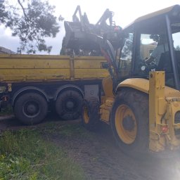 Kuba mąkosa - Żółta koparko-ładowarka Volvo BL71 ładuje gruz do wywrotki obok ogrodzenia panelowego. Widoczny operator w kabinie i fragment drzewa w tle.