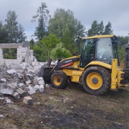 Kuba mąkosa - Żółta koparko-ładowarka Volvo obok sterty gruzu i fragmentu ściany po wyburzeniu. W tle drzewa i pochmurne niebo. Widoczny operator w kabinie maszyny.