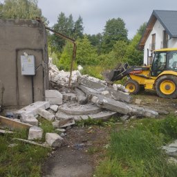 Kuba mąkosa - Żółta koparko-ładowarka Volvo BL71 na tle zniszczonej ściany budynku i nowego domu. Gruz i resztki betonu na pierwszym planie. Widoczne drzewa w tle.