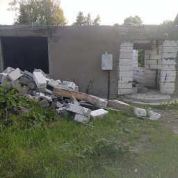 Kuba mąkosa - Budowa domu w trakcie: surowy stan z widocznymi bloczkami, stertą gruzu i skrzynką elektryczną na szarej ścianie. W tle dom jednorodzinny i zieleń.