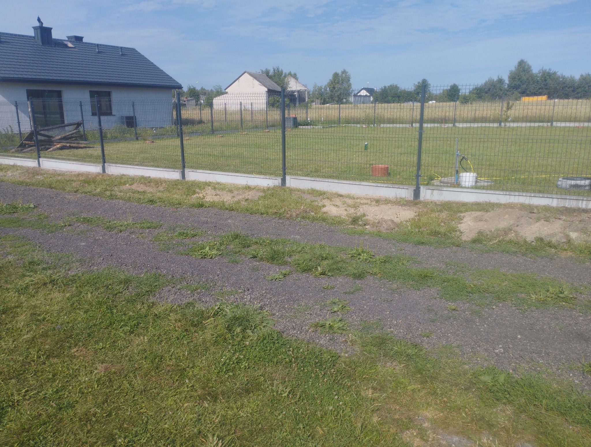 Nowoczesne ogrodzenie panelowe z podmurówką betonową na tle zieleni i zabudowań. Widoczne słupki i siatka ogrodzeniowa. Jasne, słoneczne oświetlenie.