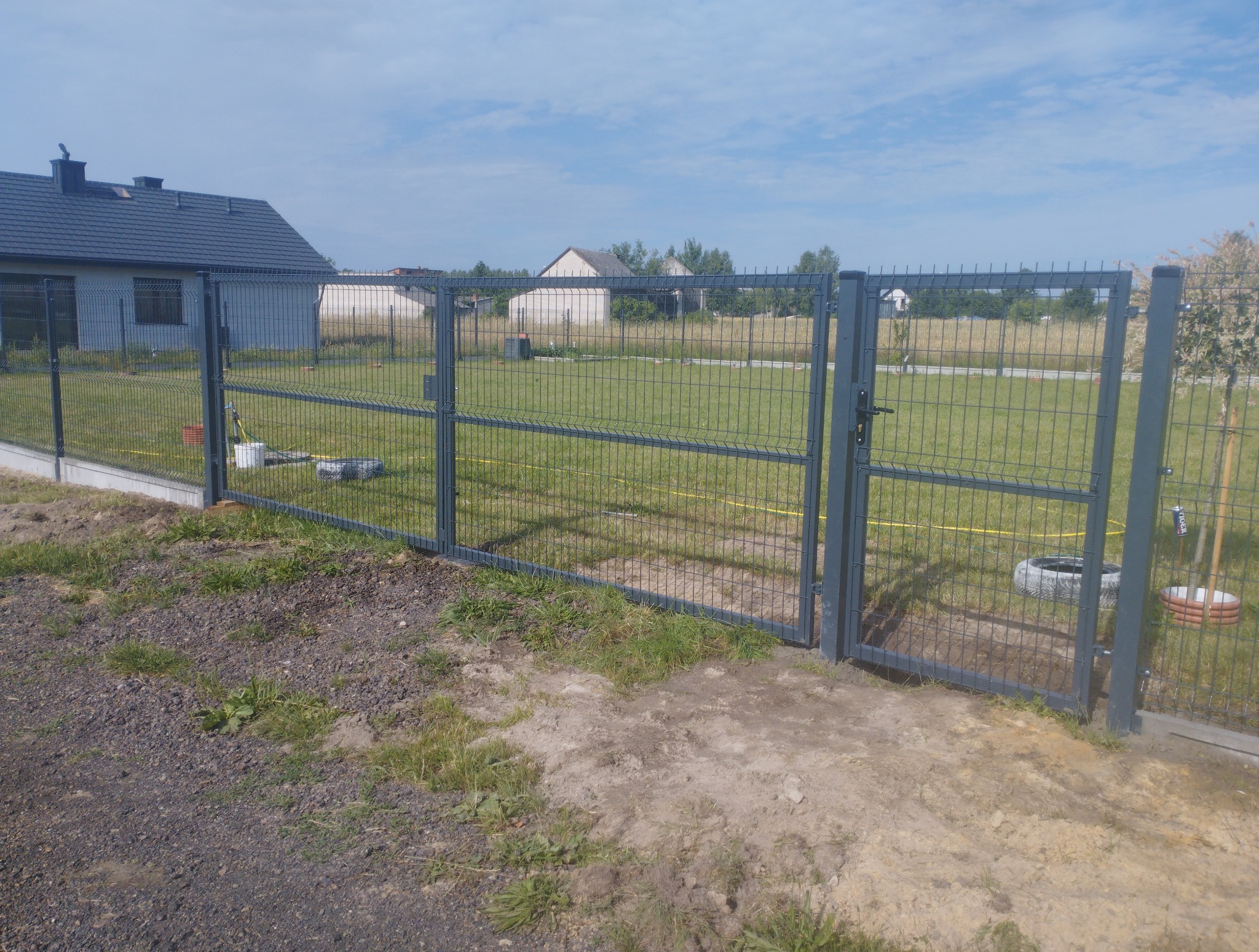 Szara, metalowa brama panelowa w Radomiu, z widoczną furtką i fragmentem ogrodzenia. W tle dom i trawnik, brama otwarta na oścież, słoneczny dzień.