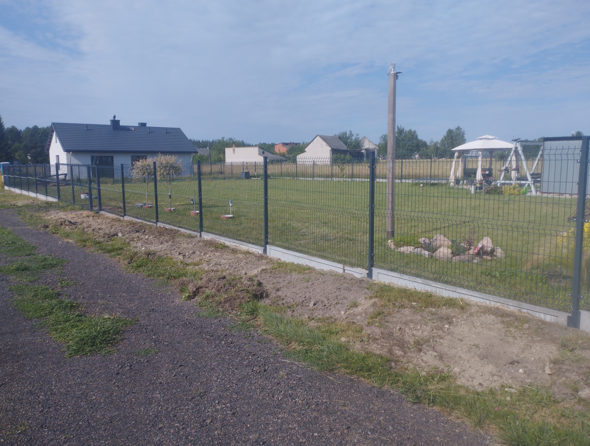 Nowoczesne ogrodzenie panelowe z podmurówką betonową, widok z boku, na tle domów jednorodzinnych i zielonego trawnika. Słoneczny dzień, błękitne niebo.