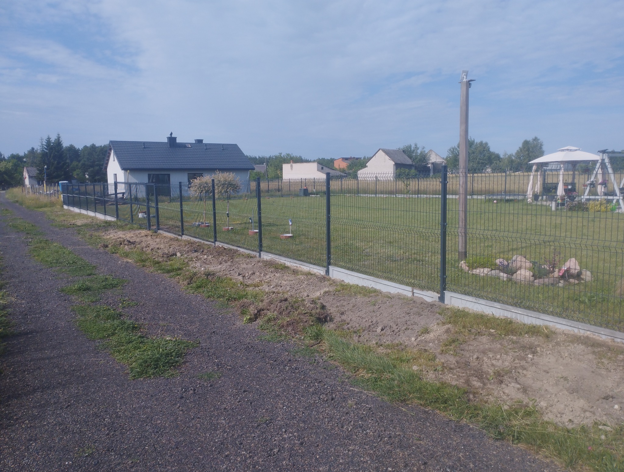 Nowo postawione ogrodzenie panelowe z podmurówką na tle wiejskiego krajobrazu z domami i zielenią. Widoczne świeżo usypane ziemią wzdłuż ogrodzenia.