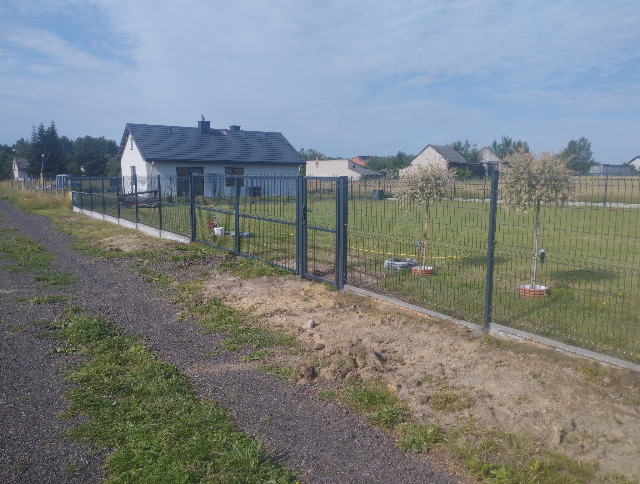 Nowoczesne ogrodzenie panelowe z bramką na betonowej podmurówce, widok z zewnątrz posesji. W tle dom jednorodzinny i drzewa.