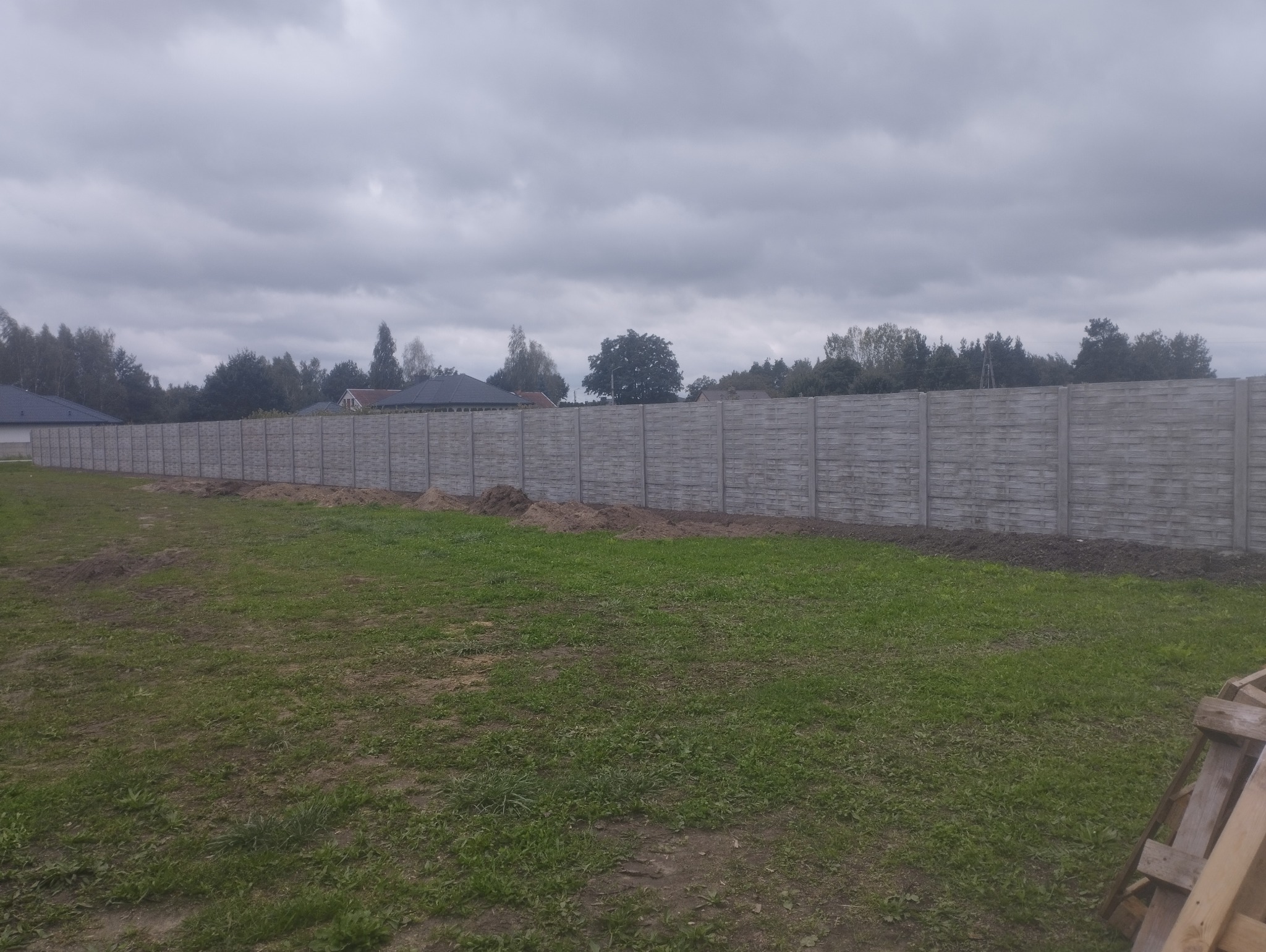 Długi, szary, prefabrykowany płot betonowy z poziomymi panelami, oddzielający posesję od drogi, na tle pochmurnego nieba i zieleni. Widoczne ślady prac ziemnych.