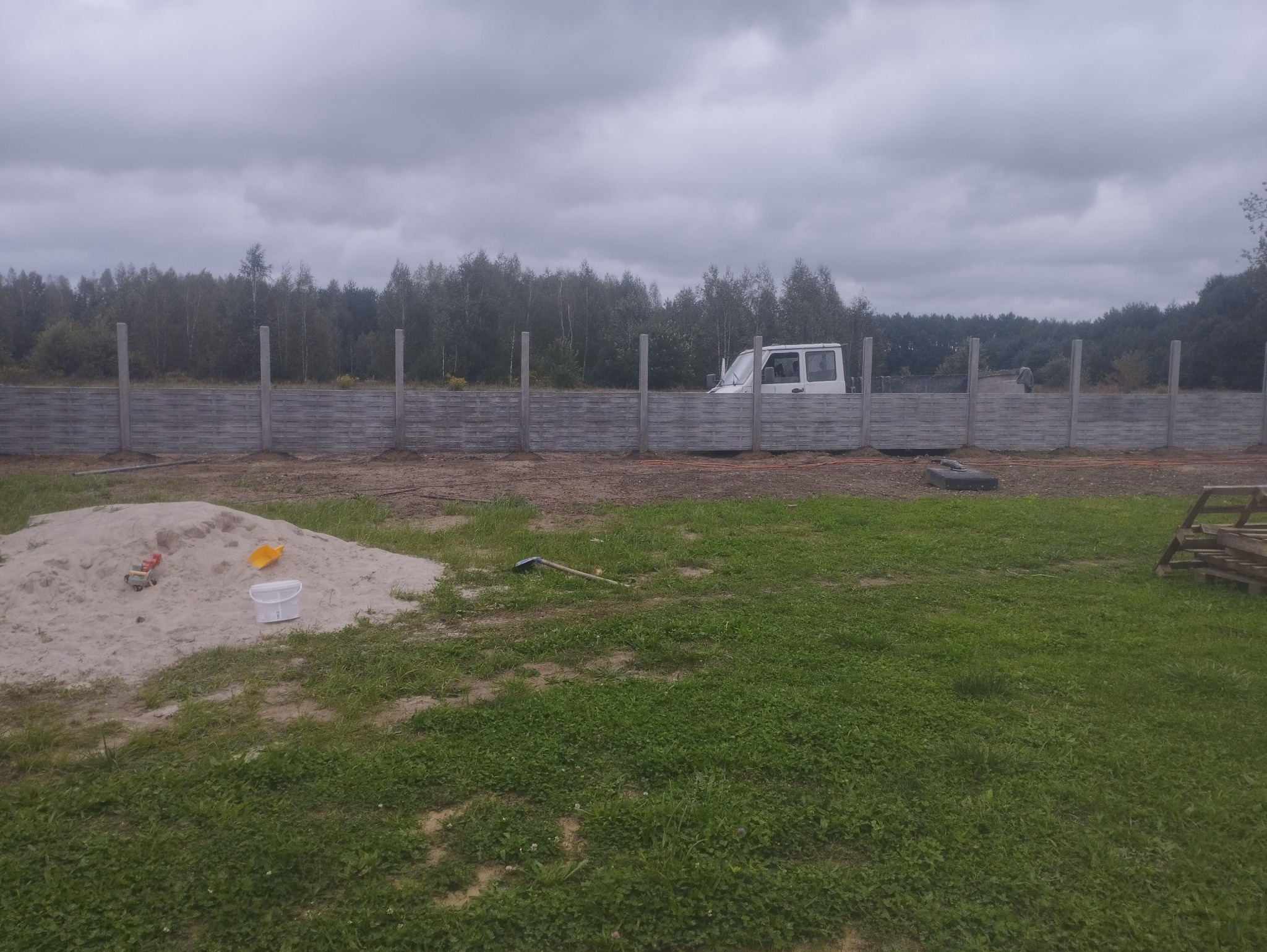 Wznoszenie ogrodzenia betonowego z prefabrykatów. Widoczne słupki i panele w trakcie montażu, sterta piasku z zabawkami i auto dostawcze w tle.