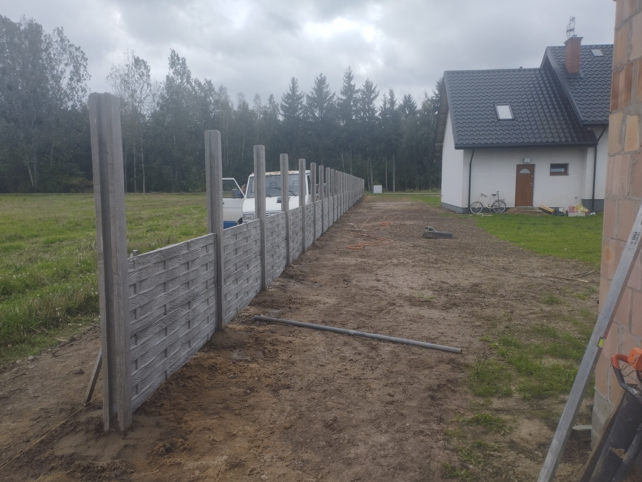 Montaż ogrodzenia betonowego imitującego drewno, widoczne słupki i panele, w tle dom jednorodzinny i las. Ujęcie z perspektywy ziemi, pochmurny dzień.