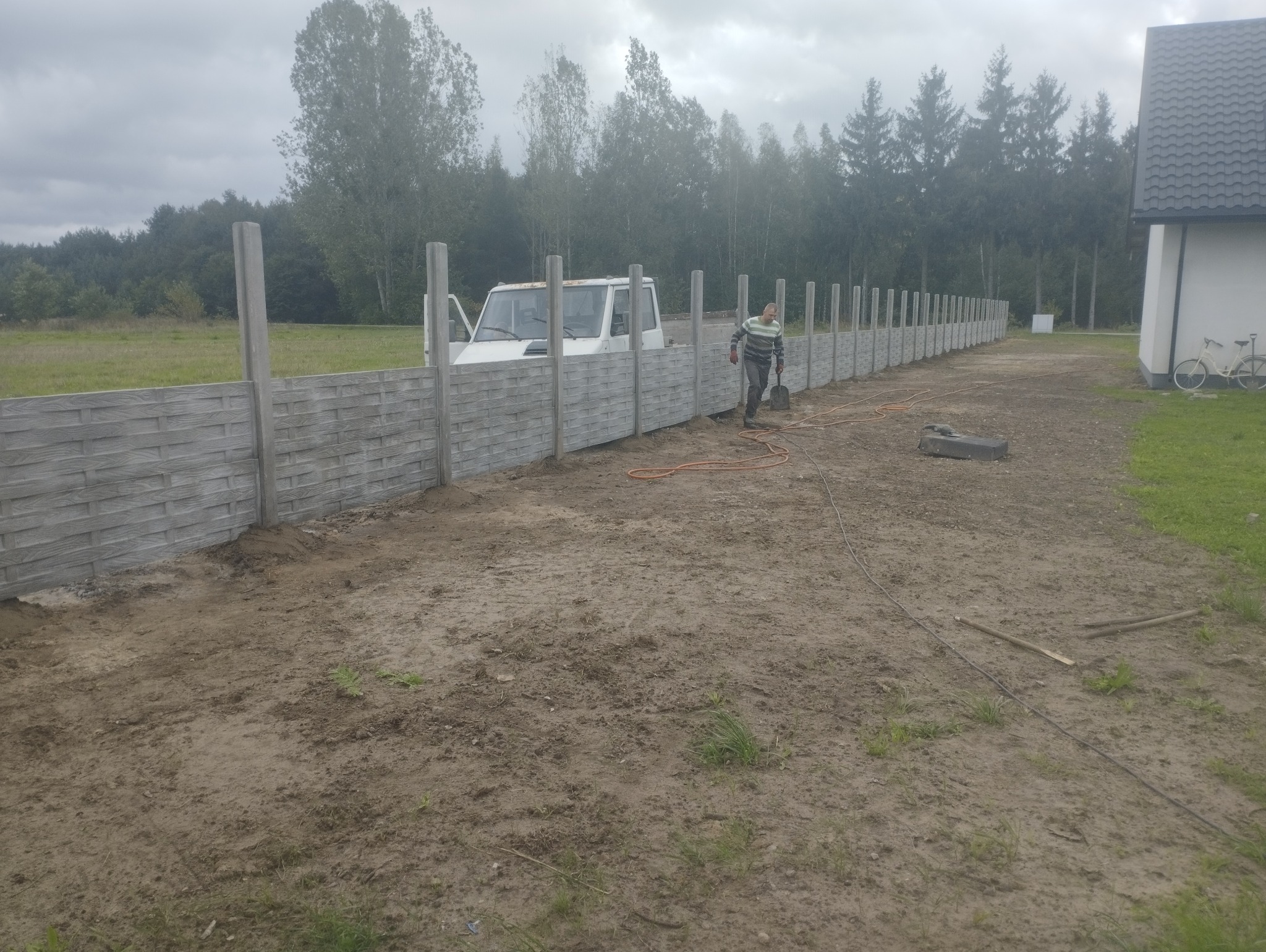 Montaż ogrodzenia betonowego z elementami dekoracyjnymi. Widoczny pracownik z łopatą, częściowo ukończony płot i samochód dostawczy w tle.