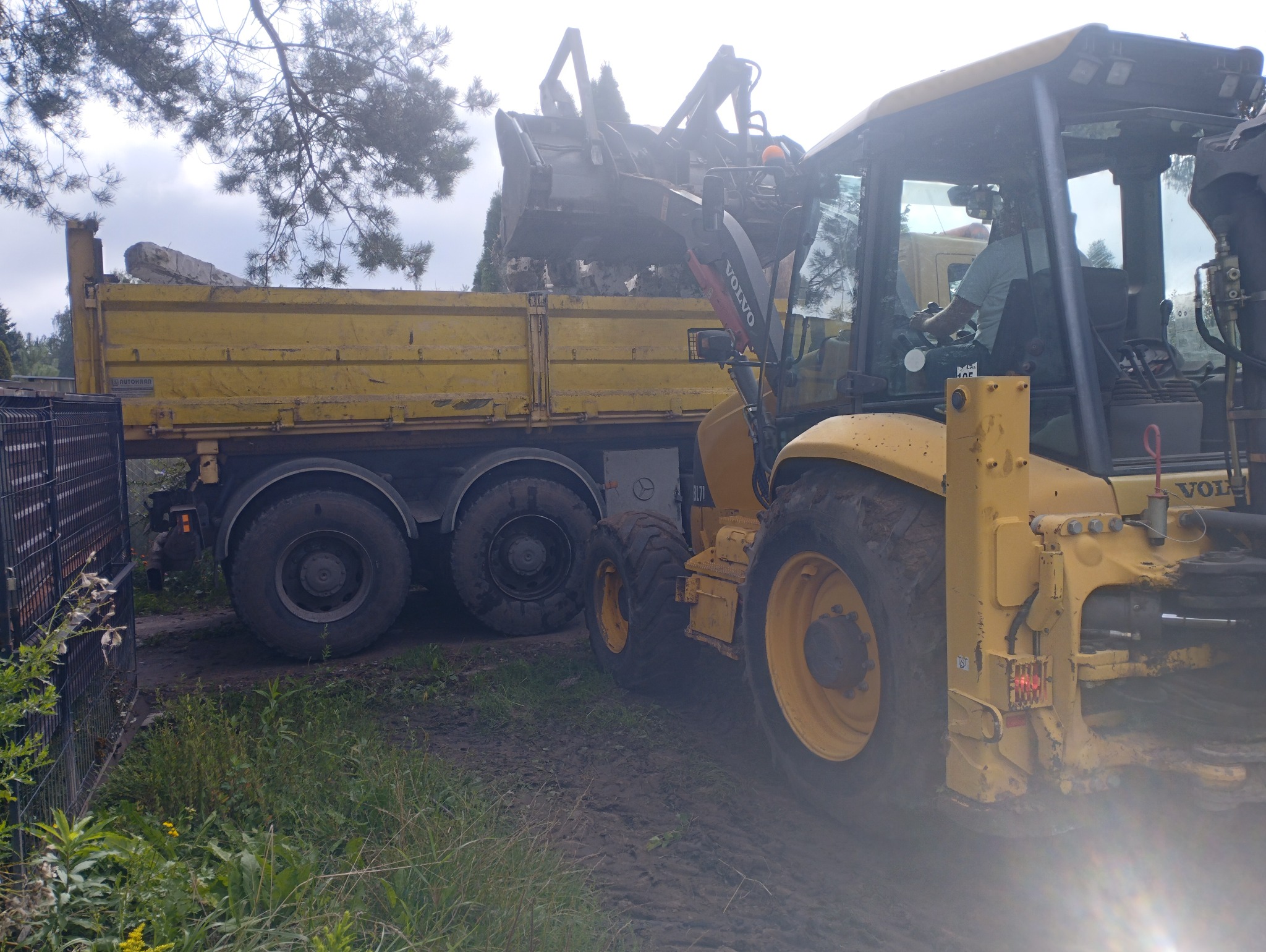 Żółta koparko-ładowarka Volvo BL71 ładuje gruz do wywrotki obok ogrodzenia panelowego. Widoczny operator w kabinie i fragment drzewa w tle.