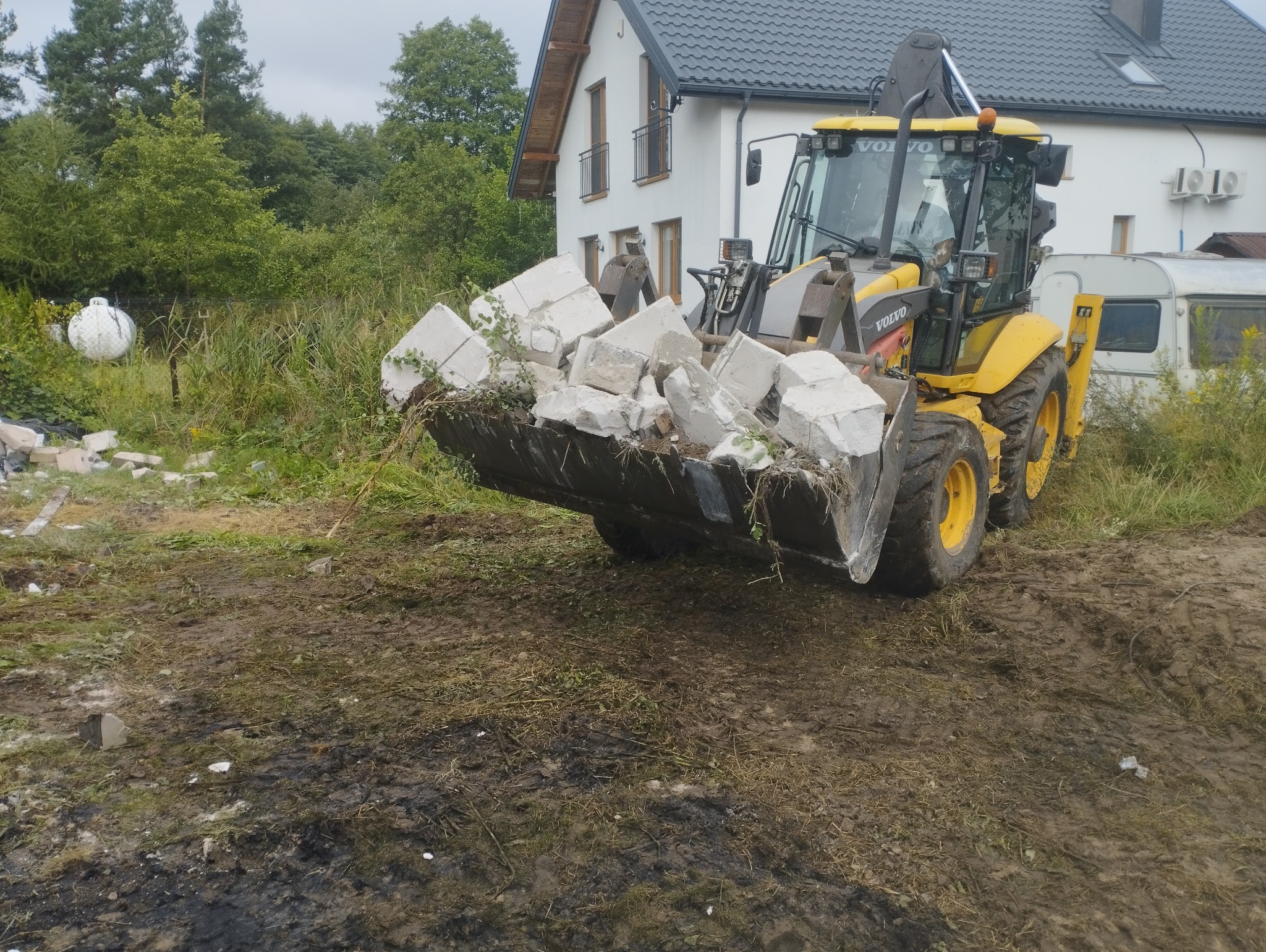 Żółta koparko-ładowarka Volvo z pełną łyżką gruzu na tle białego domu z szarą dachówką i zarośniętego terenu. Widoczne ślady opon na błotnistym podłożu.