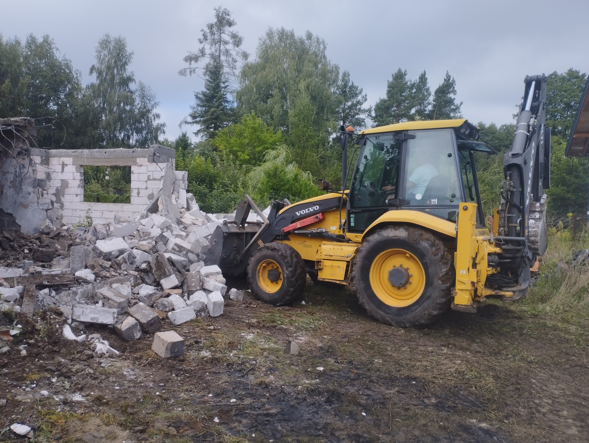 Żółta koparko-ładowarka Volvo obok sterty gruzu i fragmentu ściany po wyburzeniu. W tle drzewa i pochmurne niebo. Widoczny operator w kabinie maszyny.