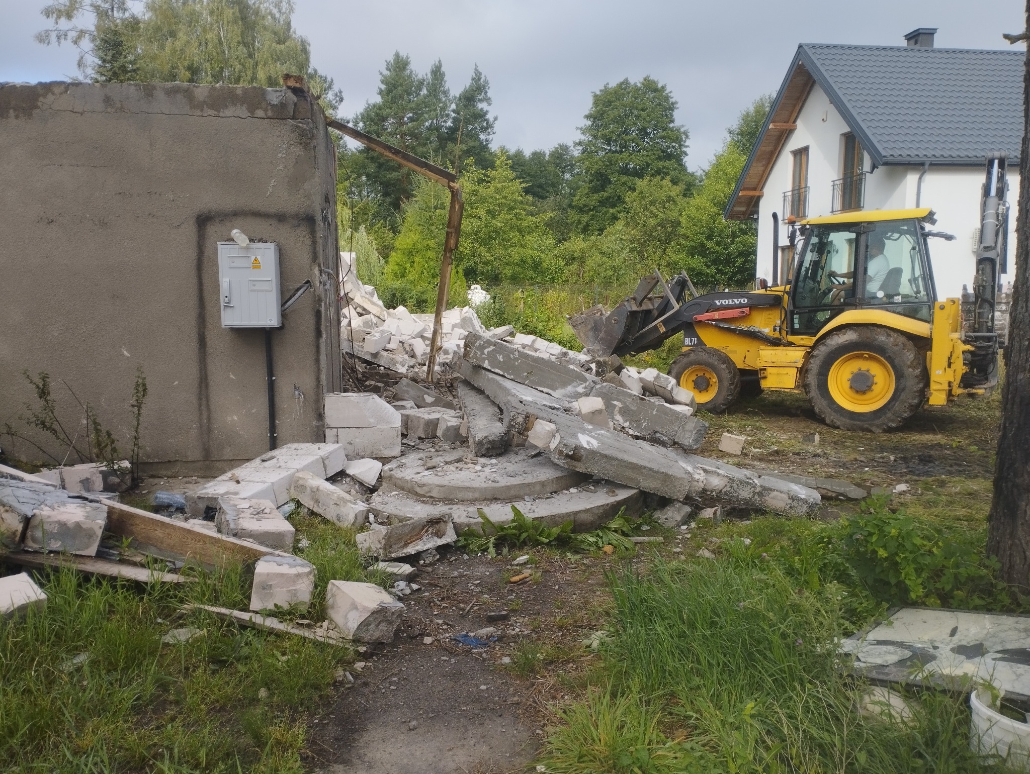 Żółta koparko-ładowarka Volvo BL71 na tle zniszczonej ściany budynku i nowego domu. Gruz i resztki betonu na pierwszym planie. Widoczne drzewa w tle.
