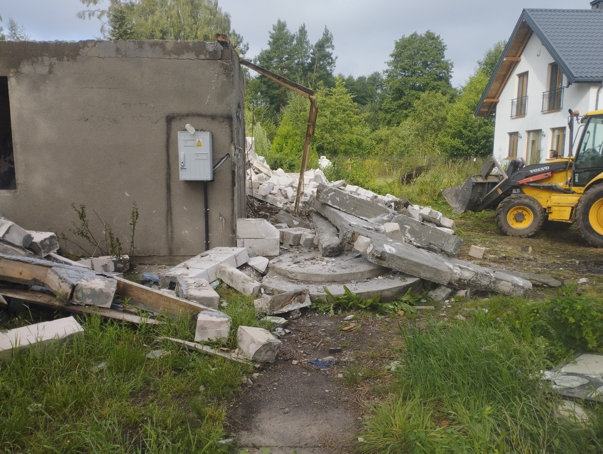 Po wyburzeniu: gruzowisko z fragmentami ścian i fundamentów, z żółtą koparką w tle, sugerujące prace rozbiórkowe w otoczeniu zieleni i domu.