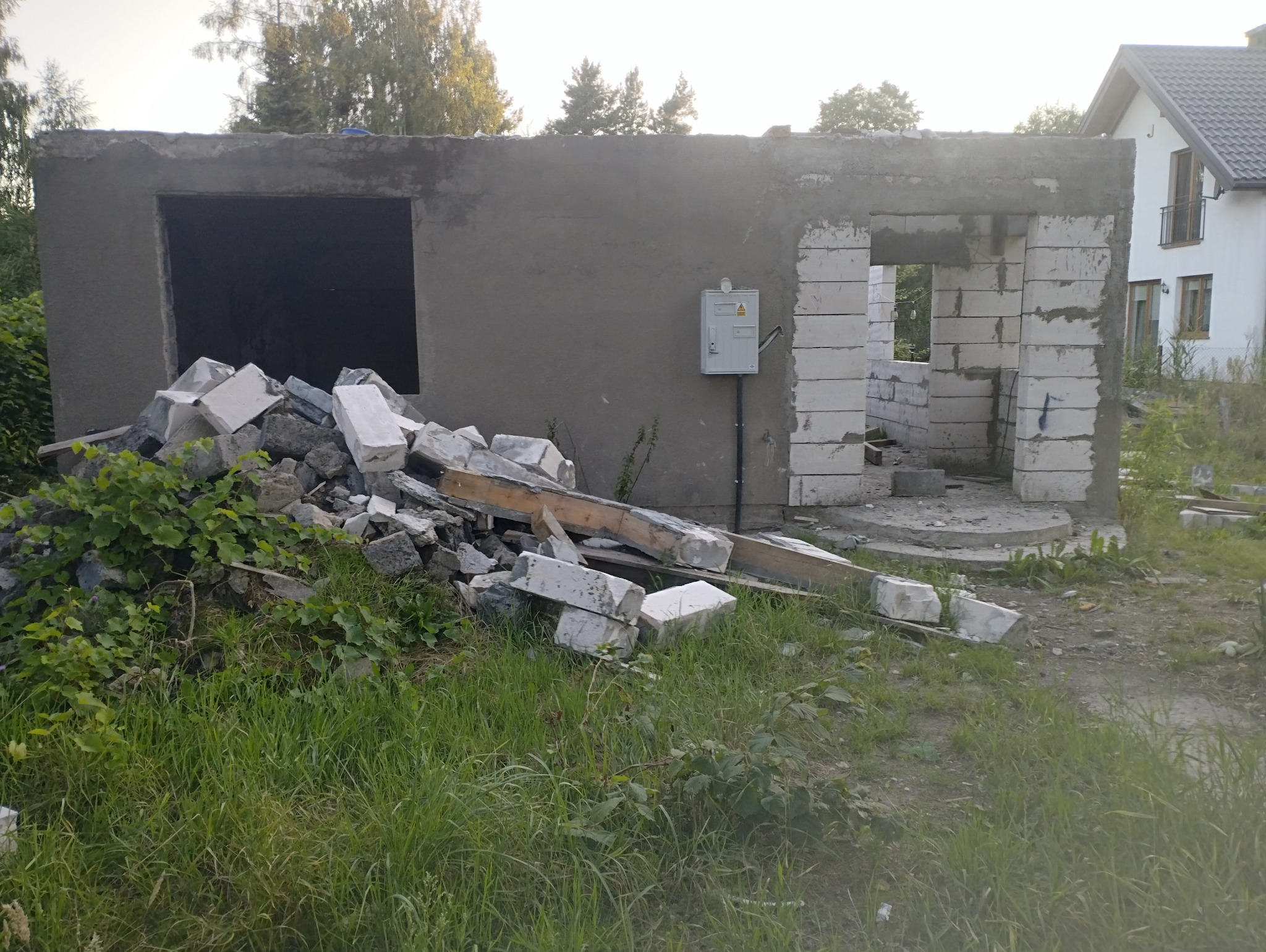 Budowa domu w trakcie: surowy stan z widocznymi bloczkami, stertą gruzu i skrzynką elektryczną na szarej ścianie. W tle dom jednorodzinny i zieleń.