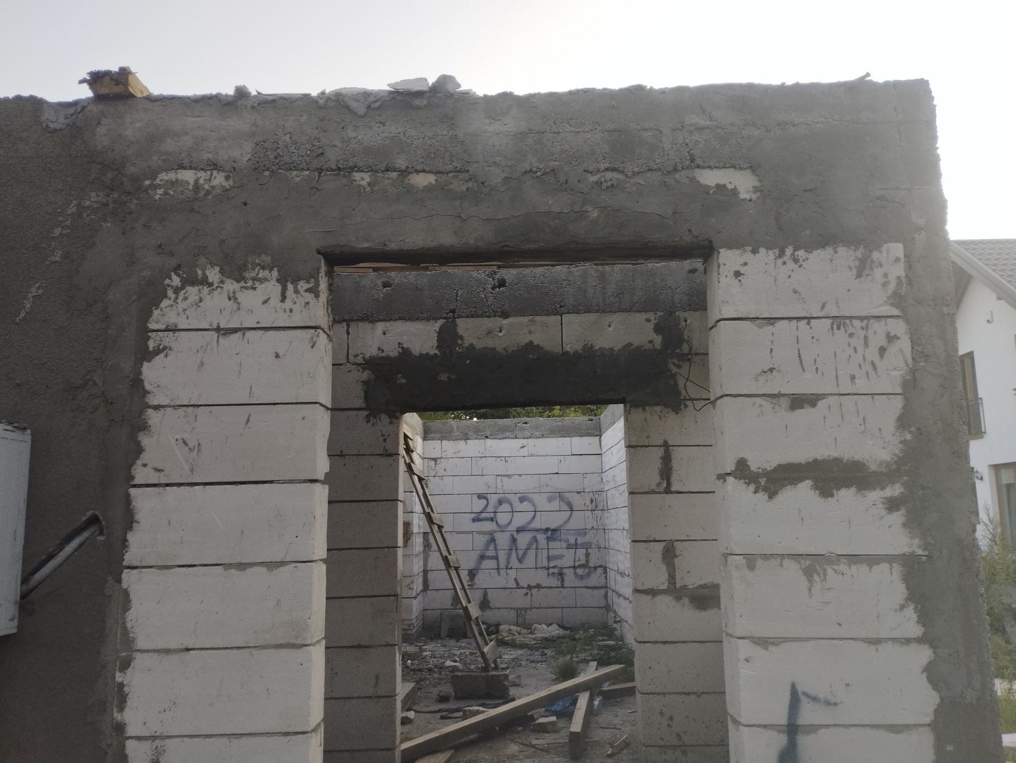 Surowy stan ściany z betonu komórkowego z widocznym nadprożem i śladami zaprawy, w tle drabina oparta o ścianę z graffiti. Etap budowy domu.