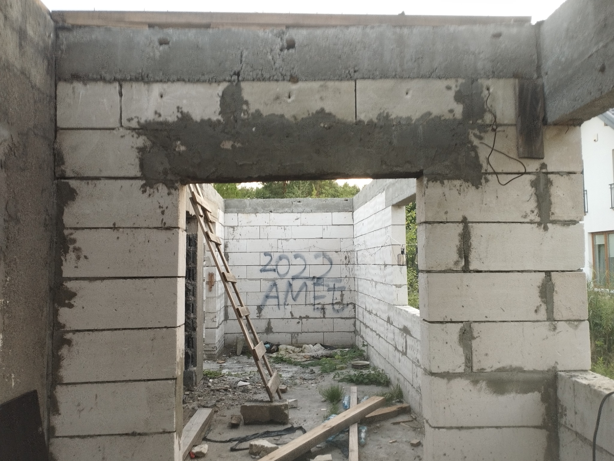 Surowy stan budynku w trakcie budowy z białych bloczków, widoczne ślady zaprawy, drabina oparta o ścianę, napis '2022 AMEL' na wewnętrznej ścianie.