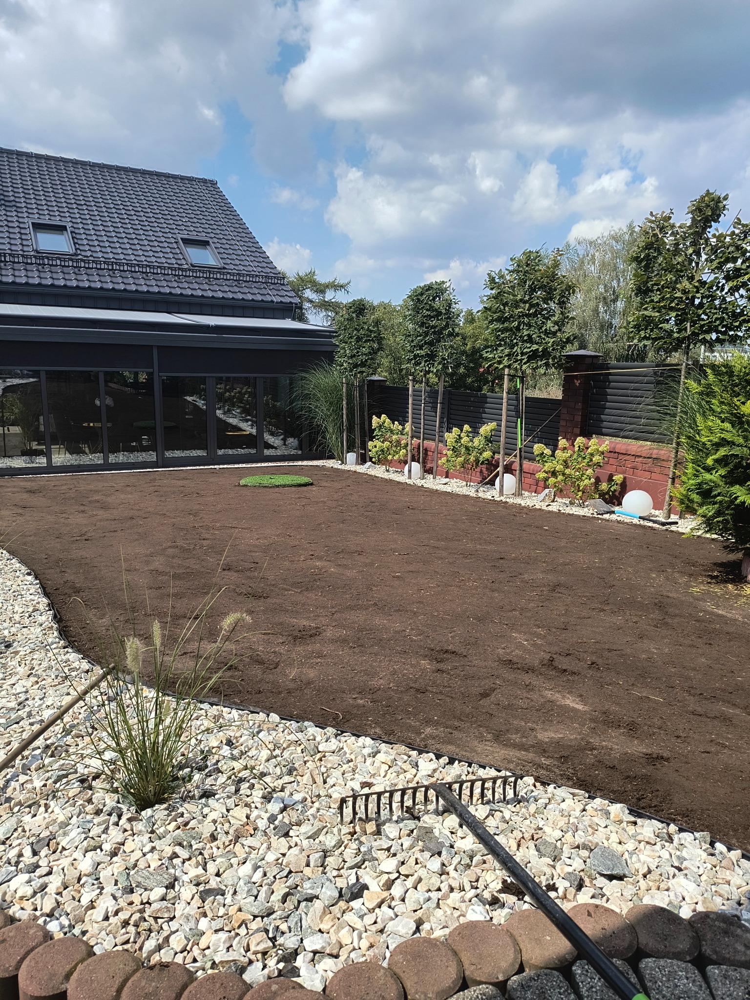 Przygotowany teren pod trawnik z rolki: wyrównana ziemia, obrzeża z kamieni, widoczny fragment trawy w rolce, grabie, dom w tle.