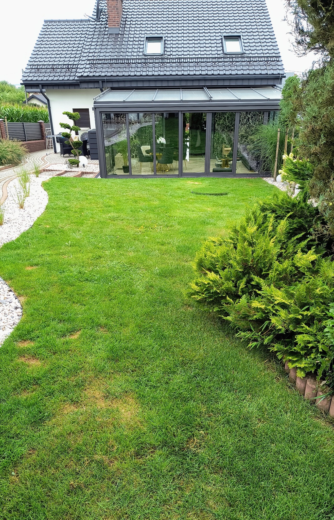 Widok na świeżo ułożony trawnik z rolki przed domem z przeszkloną werandą, z widocznymi śladami po układaniu i miejscami z nieco przeschniętą trawą.