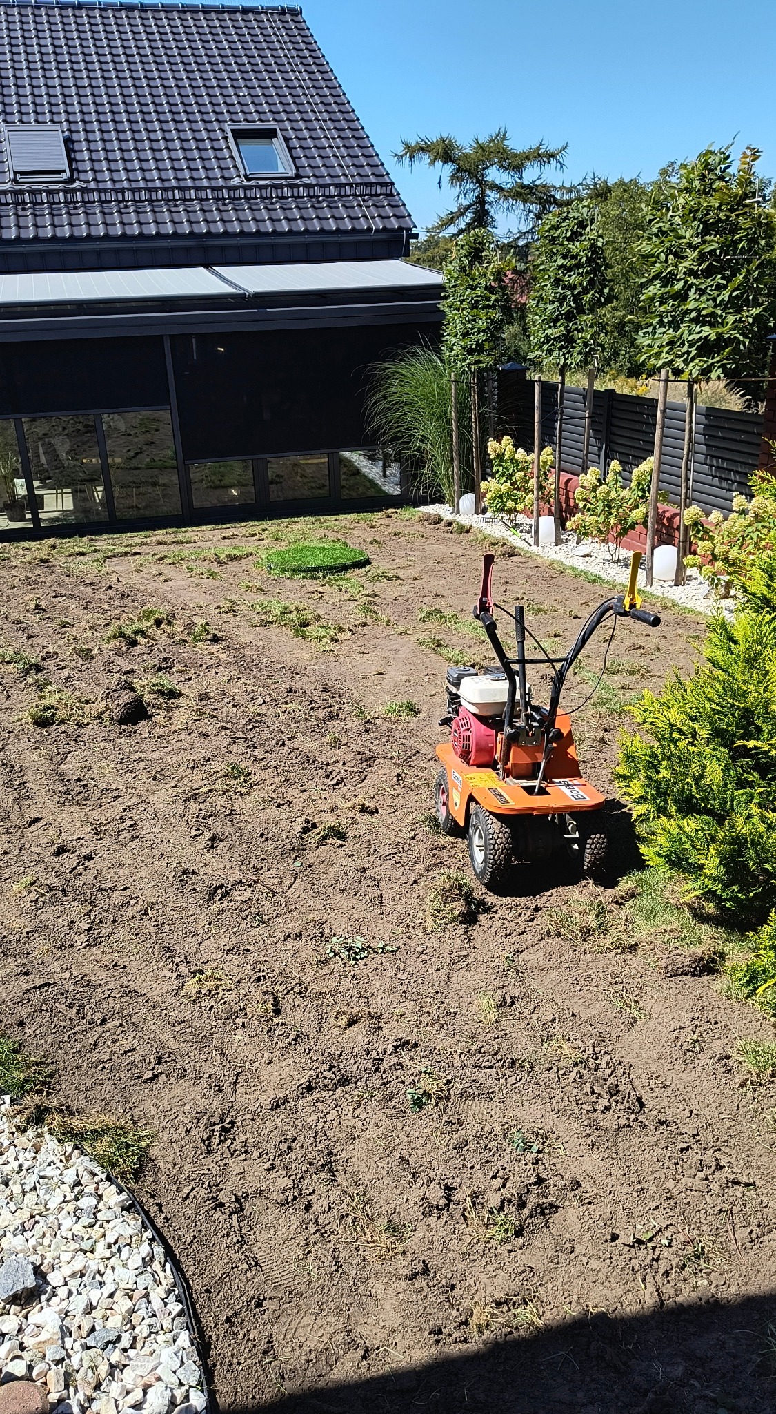 Przygotowany pod trawnik teren z glebogryzarką spalinową w ogrodzie z nowoczesnym domem w tle; widoczny fragment rolki trawy oraz obrzeże z kamyków.