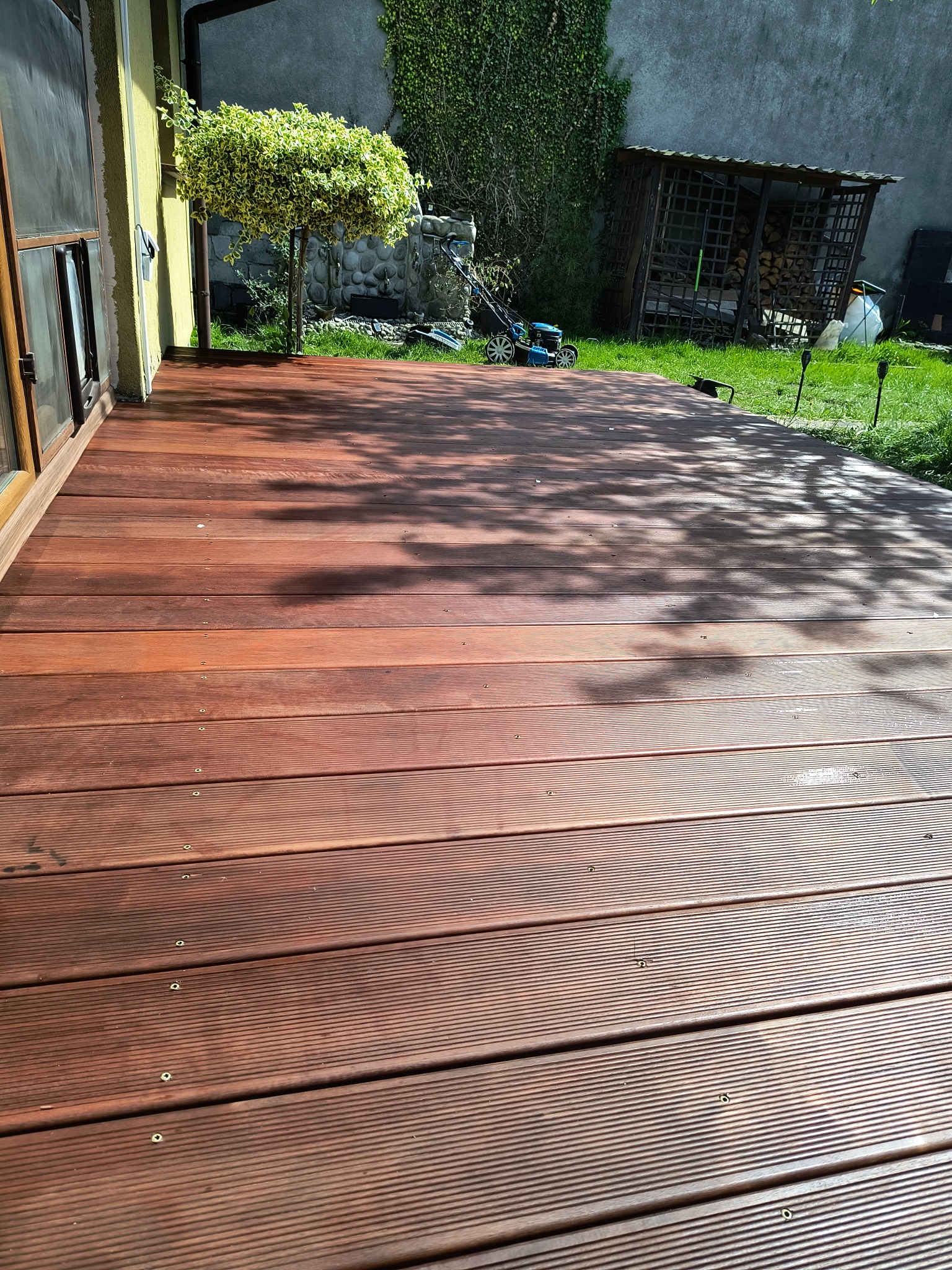 Nowy taras z desek ryflowanych massaranduba, widok z góry, z elementami ogrodu: trawnik, kosiarka, drzewko i drewutnia w tle. Ciepłe, naturalne oświetlenie.