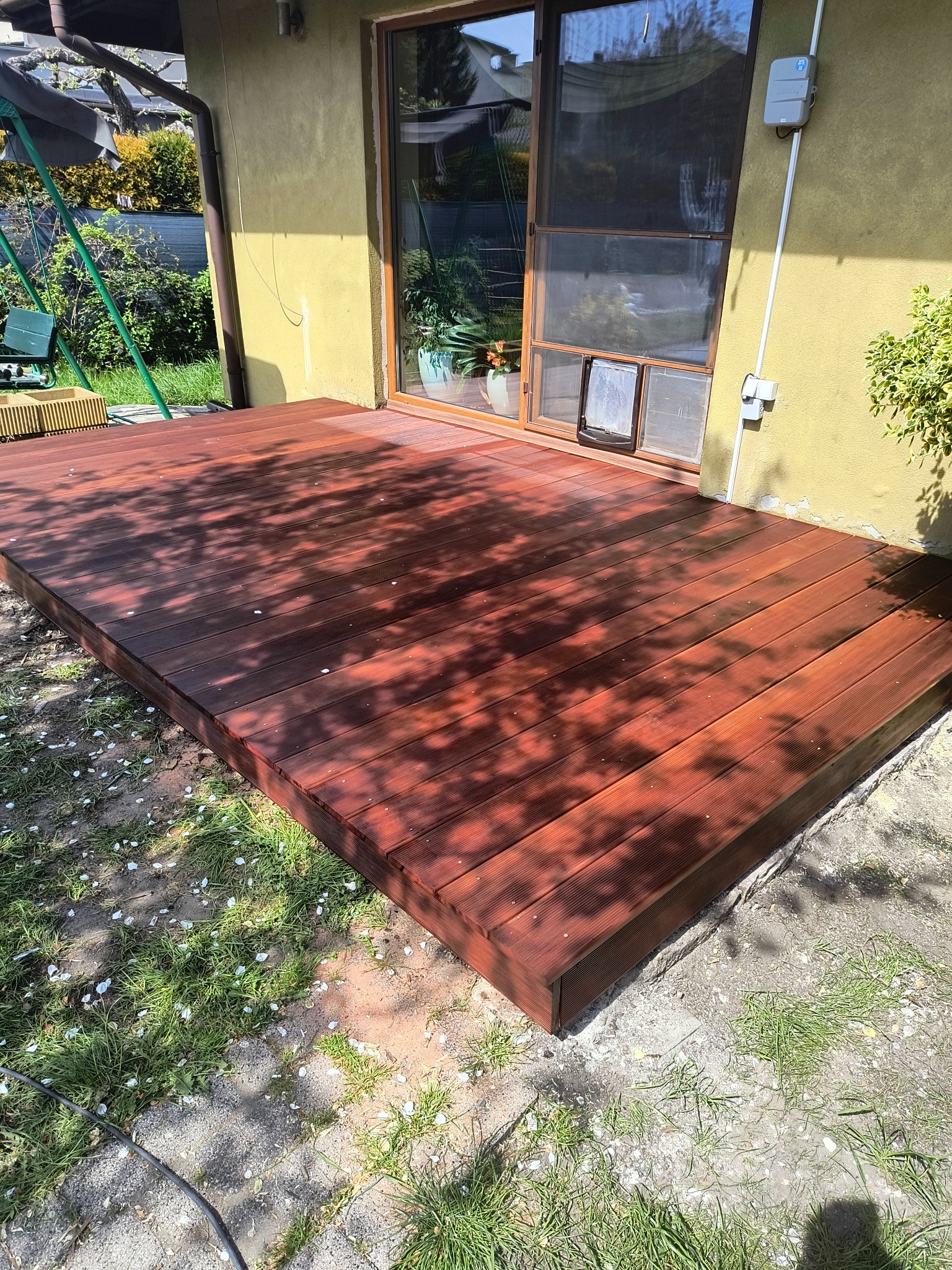 Nowy, drewniany taras z desek massaranduba, z widocznymi ryflowaniami, przylegający do żółtej ściany domu z oknem i drzwiami dla zwierząt. Widoczne cienie drzew i kwiaty na trawie.