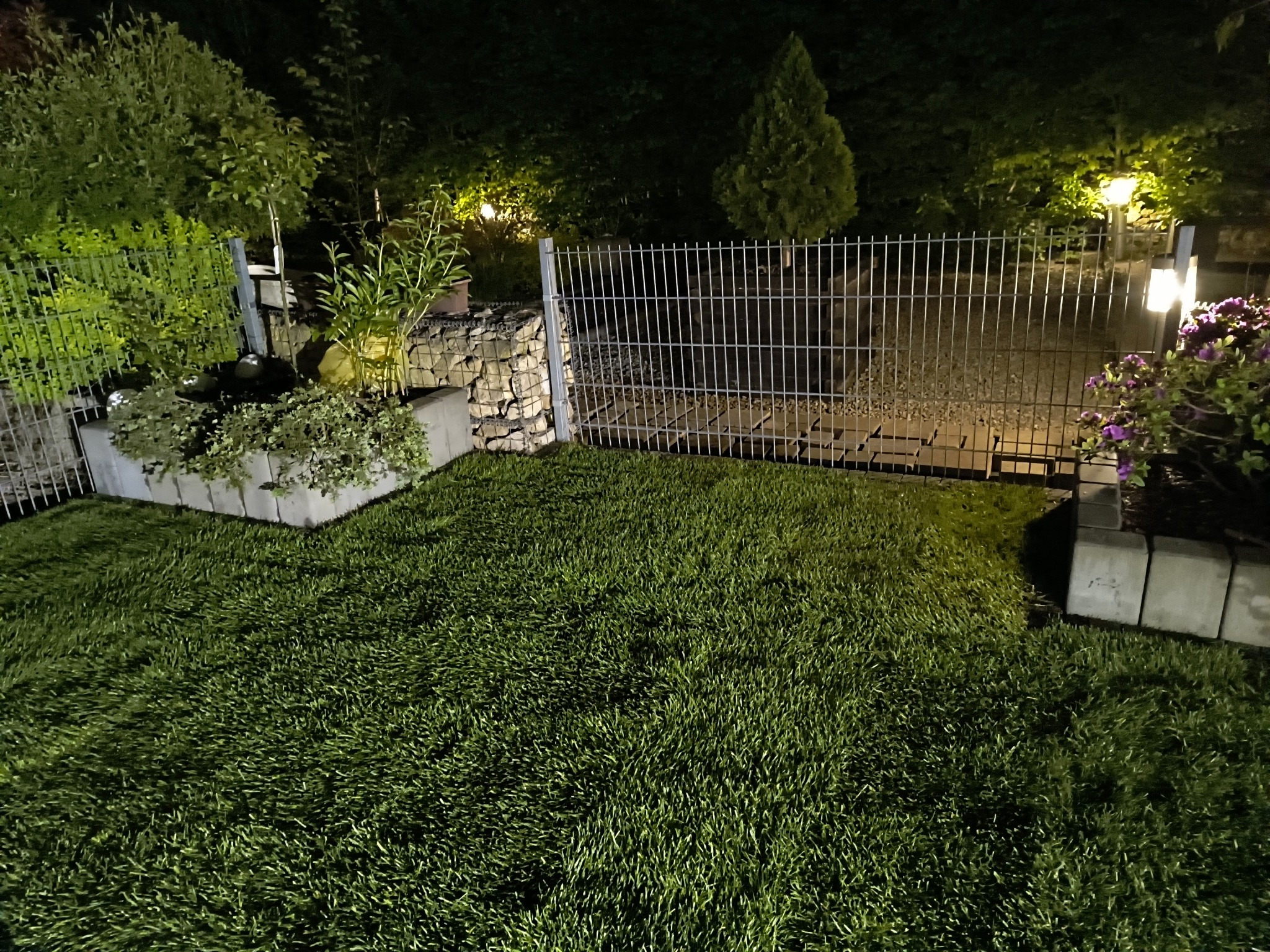 Oświetlony ogród w Krzeszowicach nocą. Widok na świeżą trawę, metalowe ogrodzenie i roślinność w donicach. Kamienne elementy dekoracyjne.